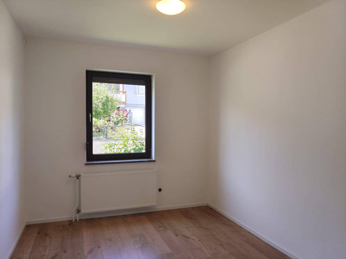 Leeres Zimmer mit Holzboden, weißen Wänden und Fenster mit Blick auf den Garten.
