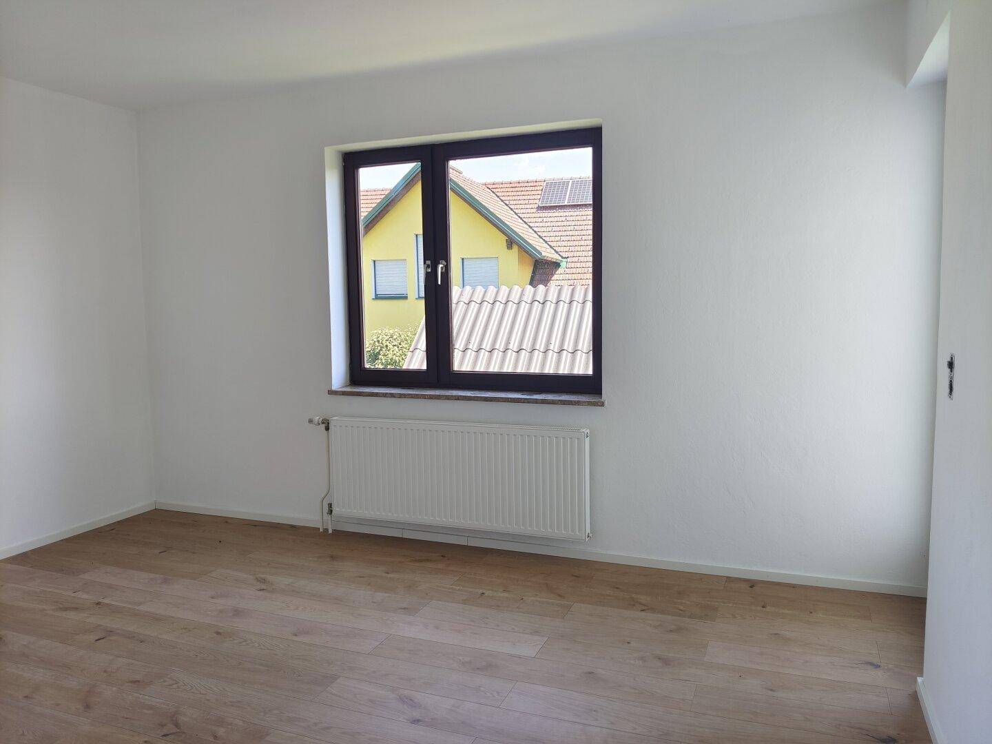 Helles, leeres Zimmer mit Holzboden und Fenster mit Blick auf ein benachbartes Haus.