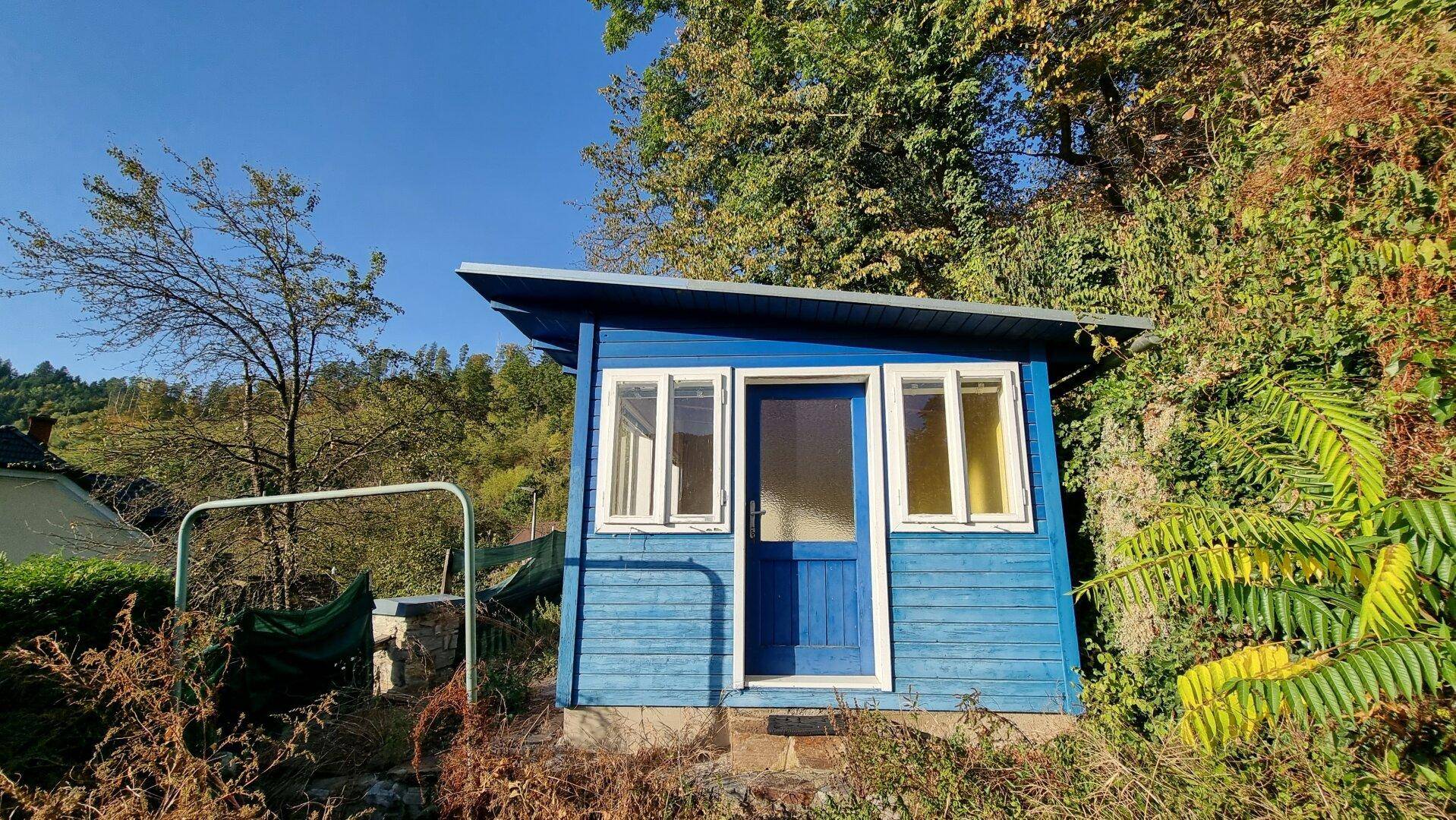 Kleine blaue Gartenhütte aus Holz, umgeben von üppigem Grün und Bäumen.