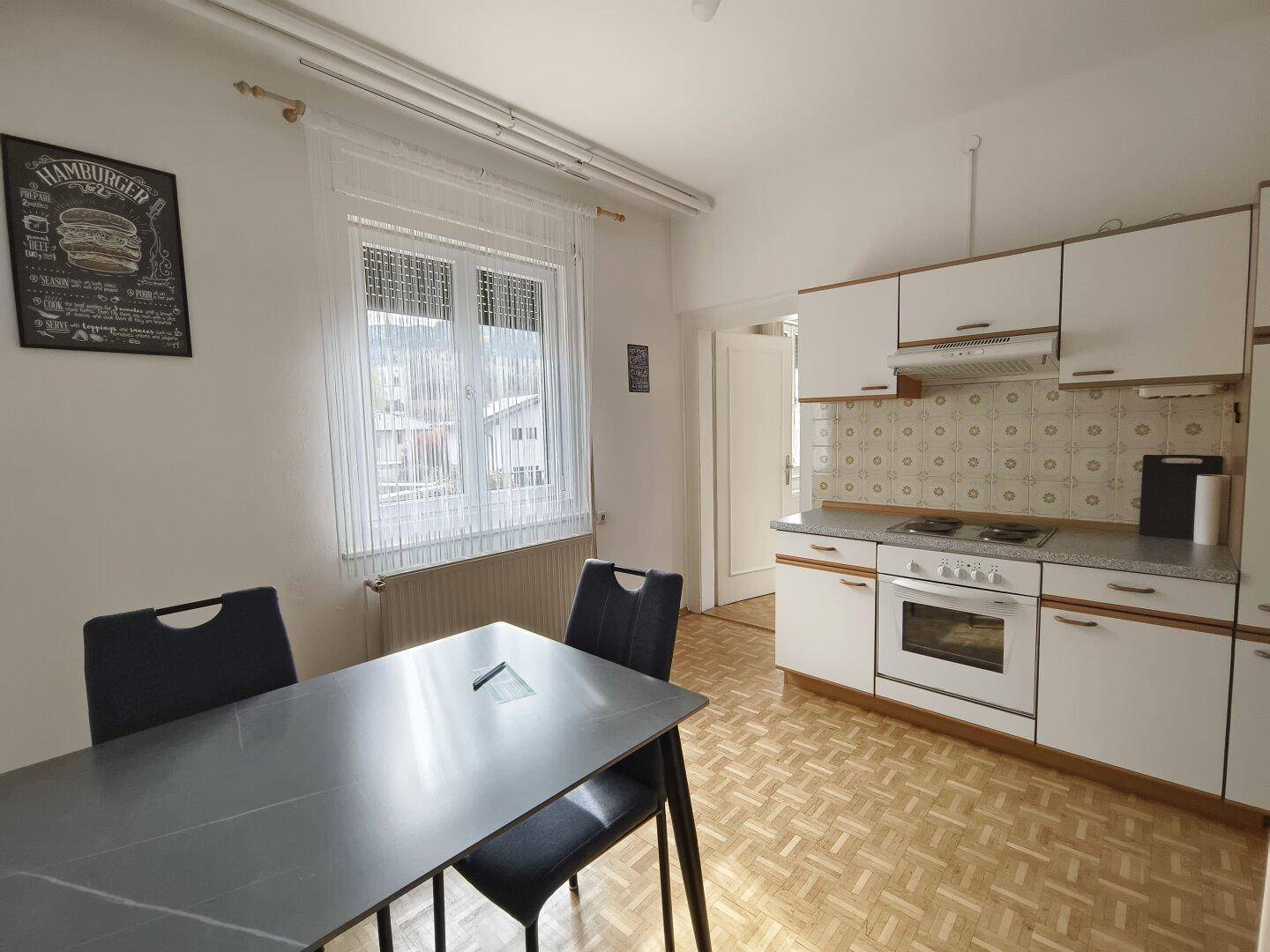 Küche mit weißen Schränken, einem Elektroherd und einem Esstisch am Fenster.