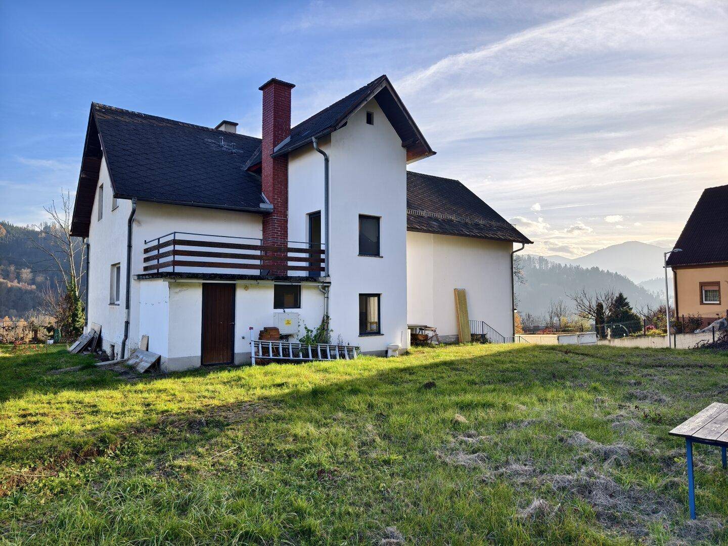 Haus mit Balkon und weitläufigem Garten, umgeben von einer hügeligen Landschaft unter blauem Himmel.