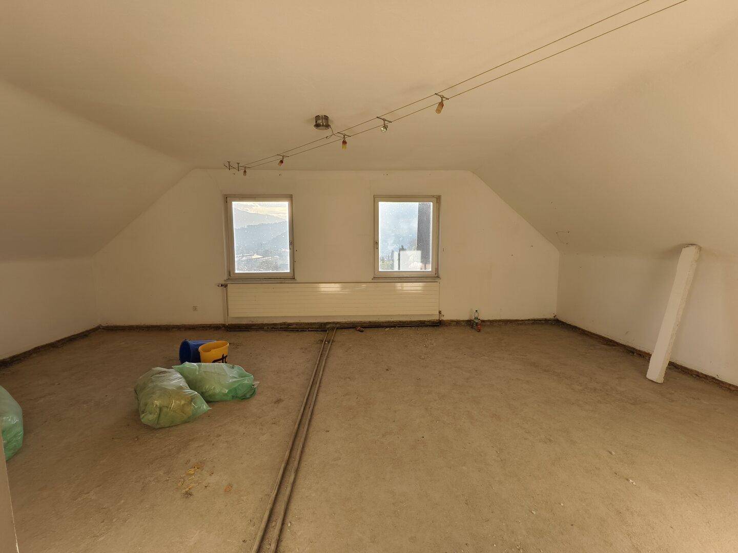 Dachgeschossraum im Rohzustand mit schrägen Wänden und Fenstern, bereit für den Ausbau.