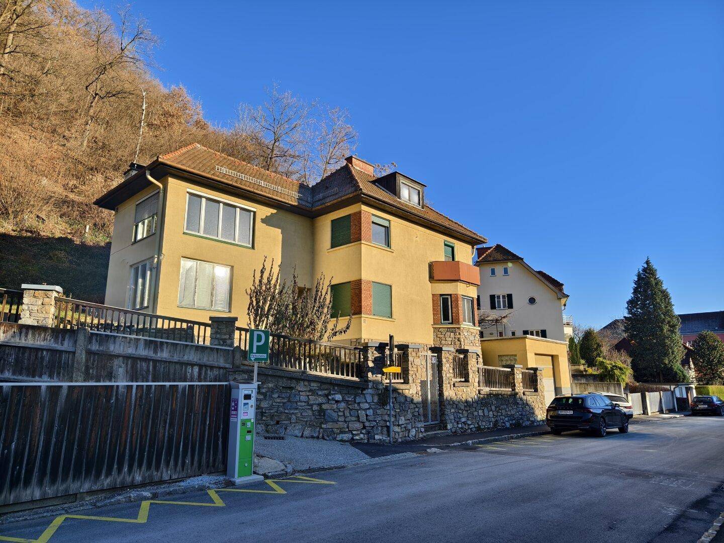 Gelbes Gebäude mit Ziegeldach und Steinsockel an einer Straße bei Sonnenschein.