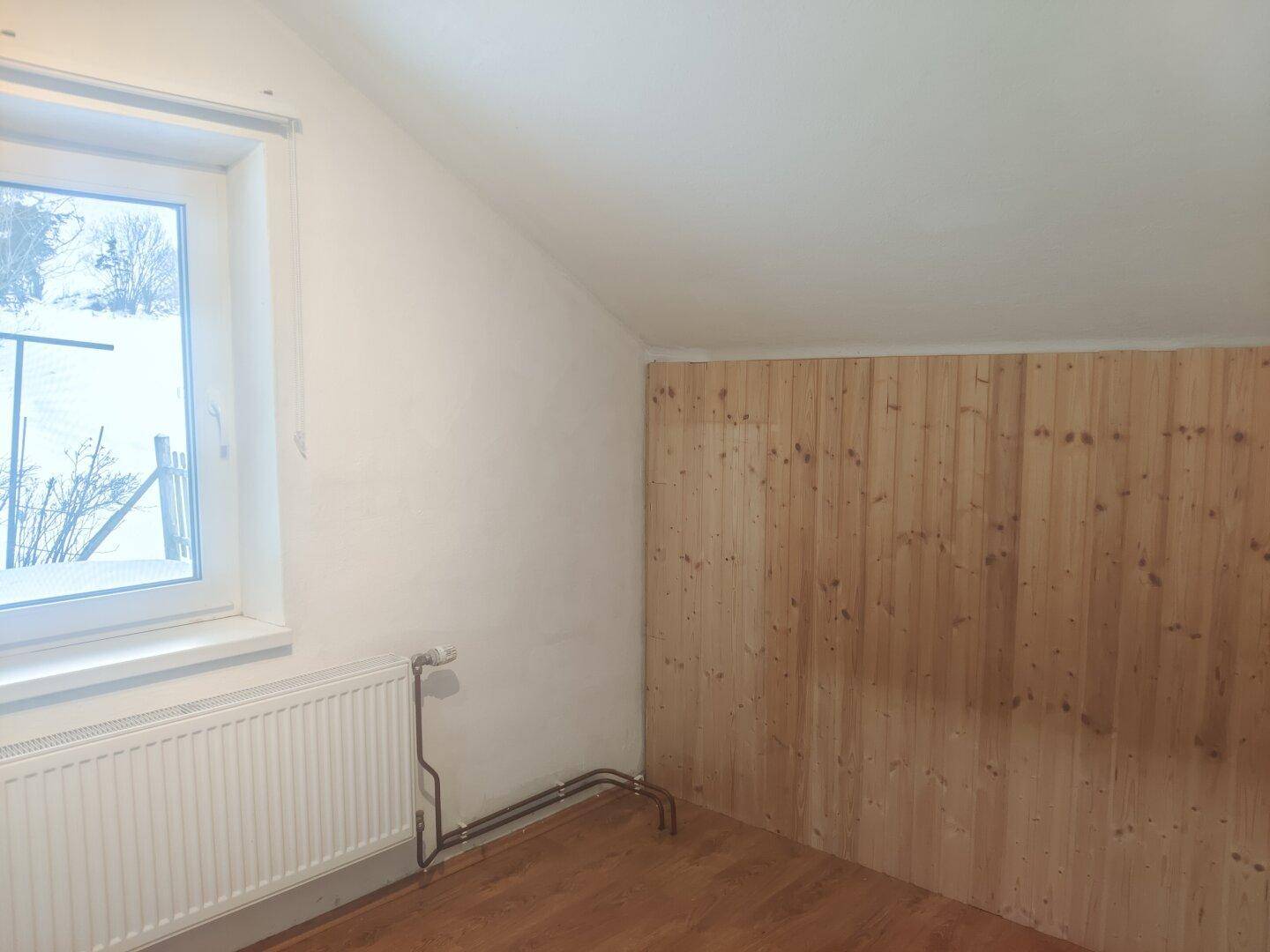 Helles Dachzimmer mit Schrägdecke, Fenster und Holzverkleidung an einer Wand.