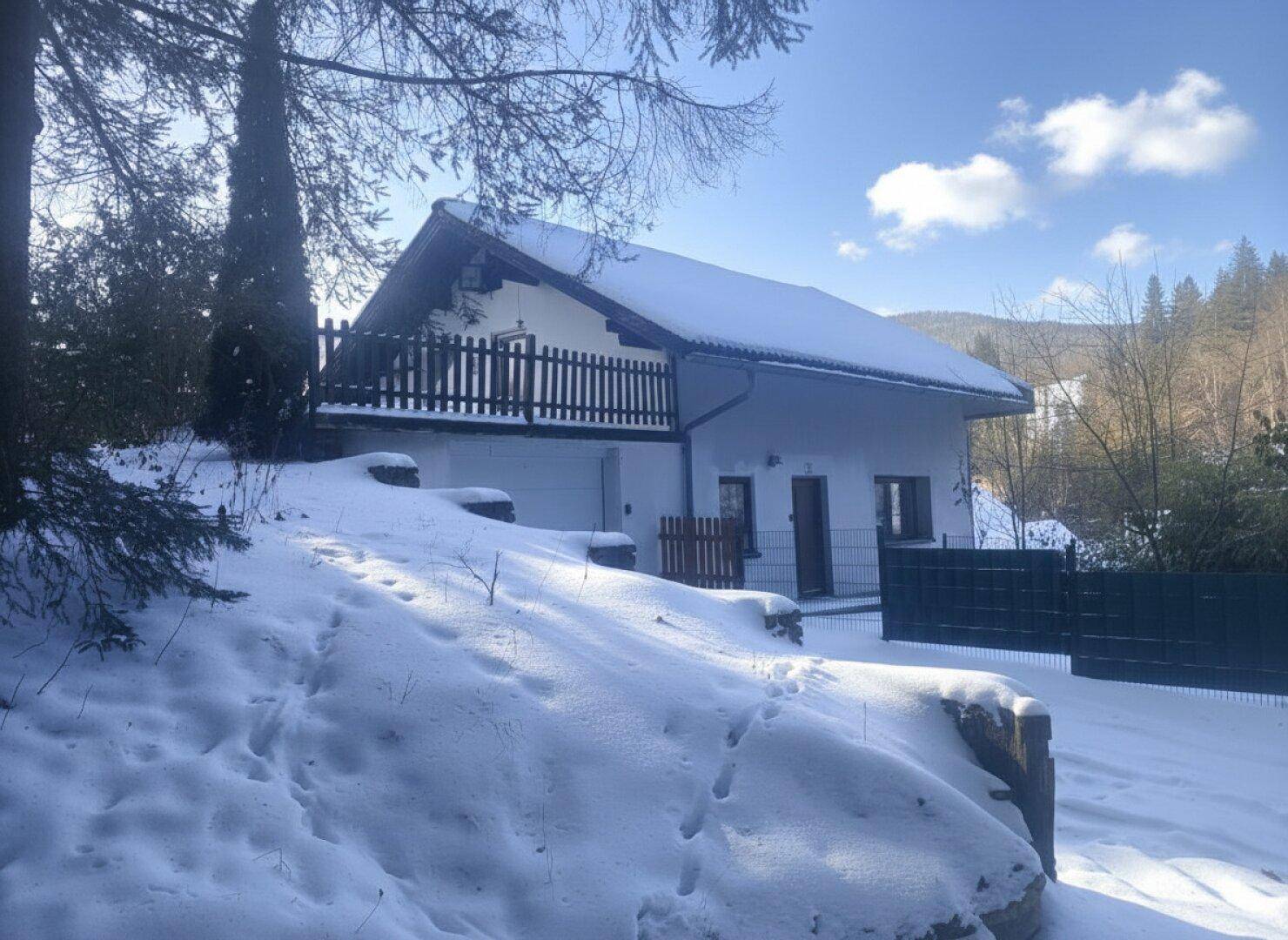 Hausfassade im Winter mit Schnee bedecktem Dach und umliegender Natur, umgeben von Bäumen.