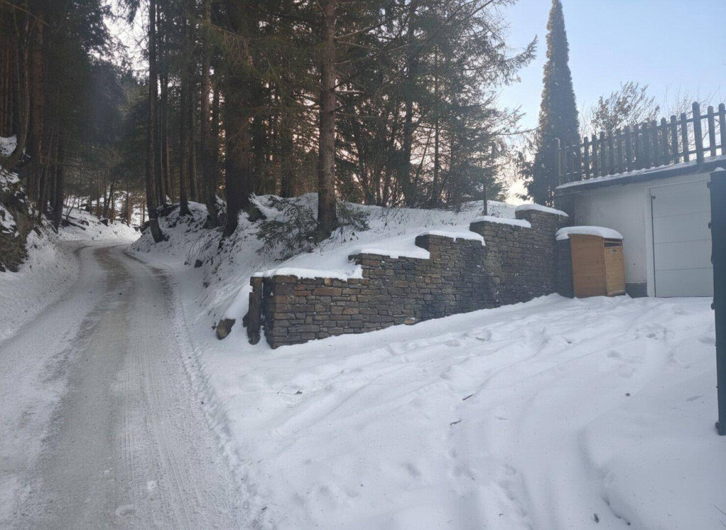 Verschneite Zufahrt mit einer Natursteinmauer und einem Gebäude im ländlichen Winterambiente.