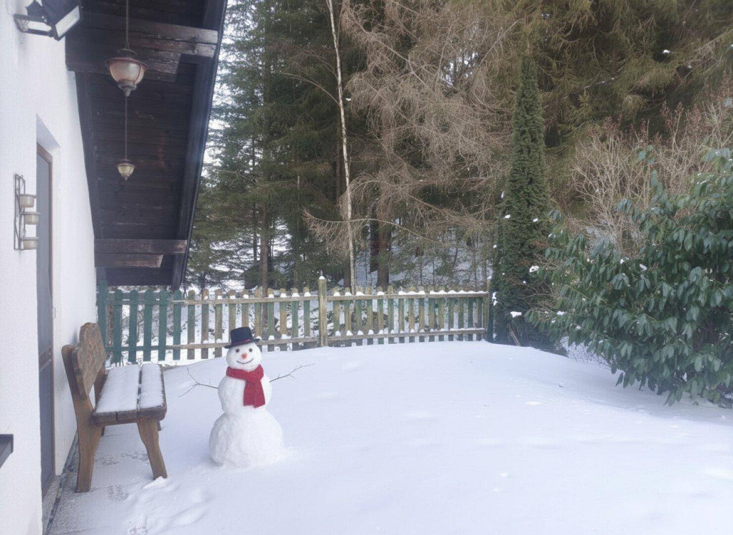 Winterliche Außenszene mit einem Schneemann neben einer Holzbank und einem Zaun im Schnee.