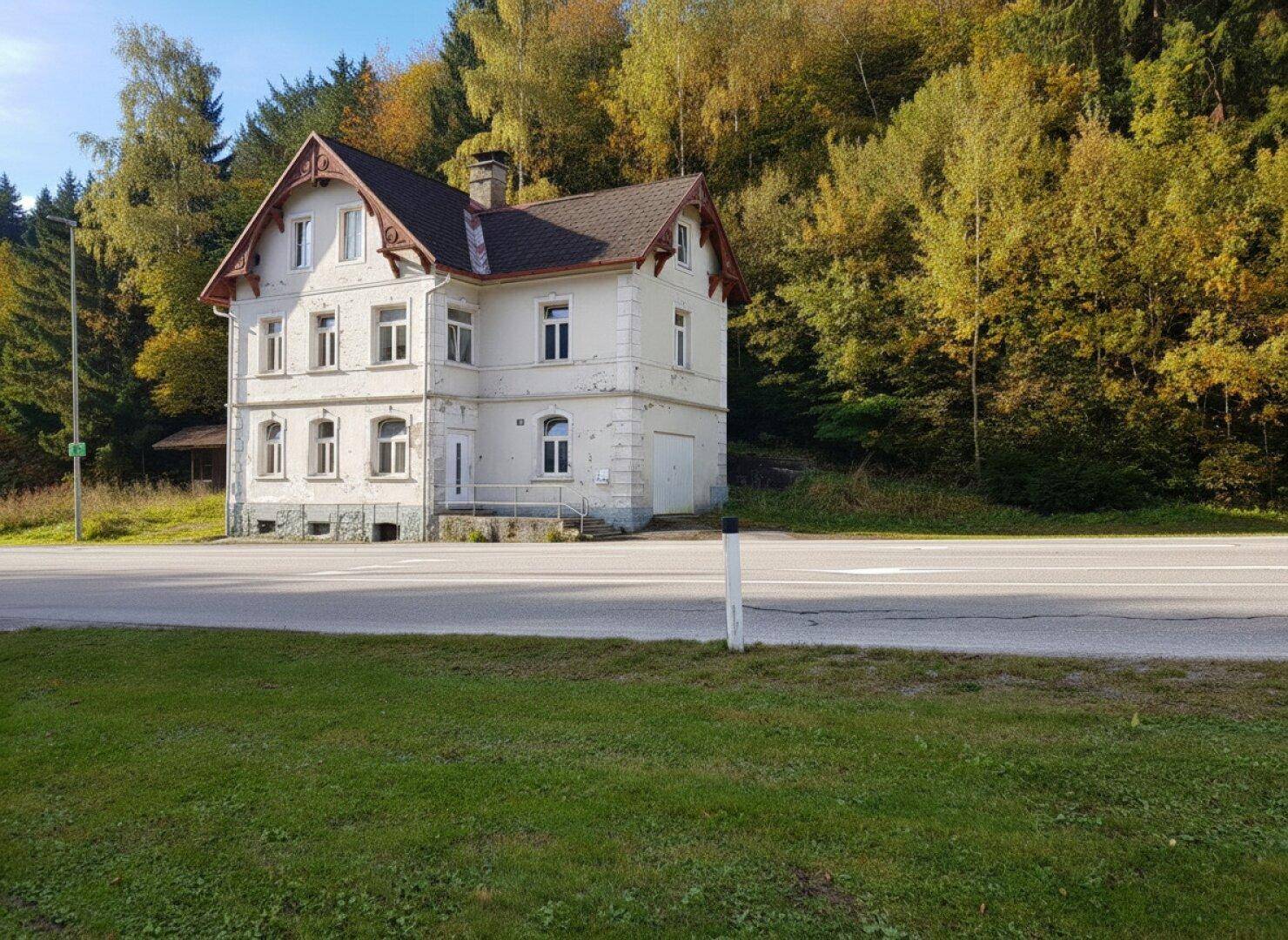 Historisches Gebäude mit weißer Fassade und rotem Dach, gelegen an einer Landstraße mit Wald im Hintergrund.