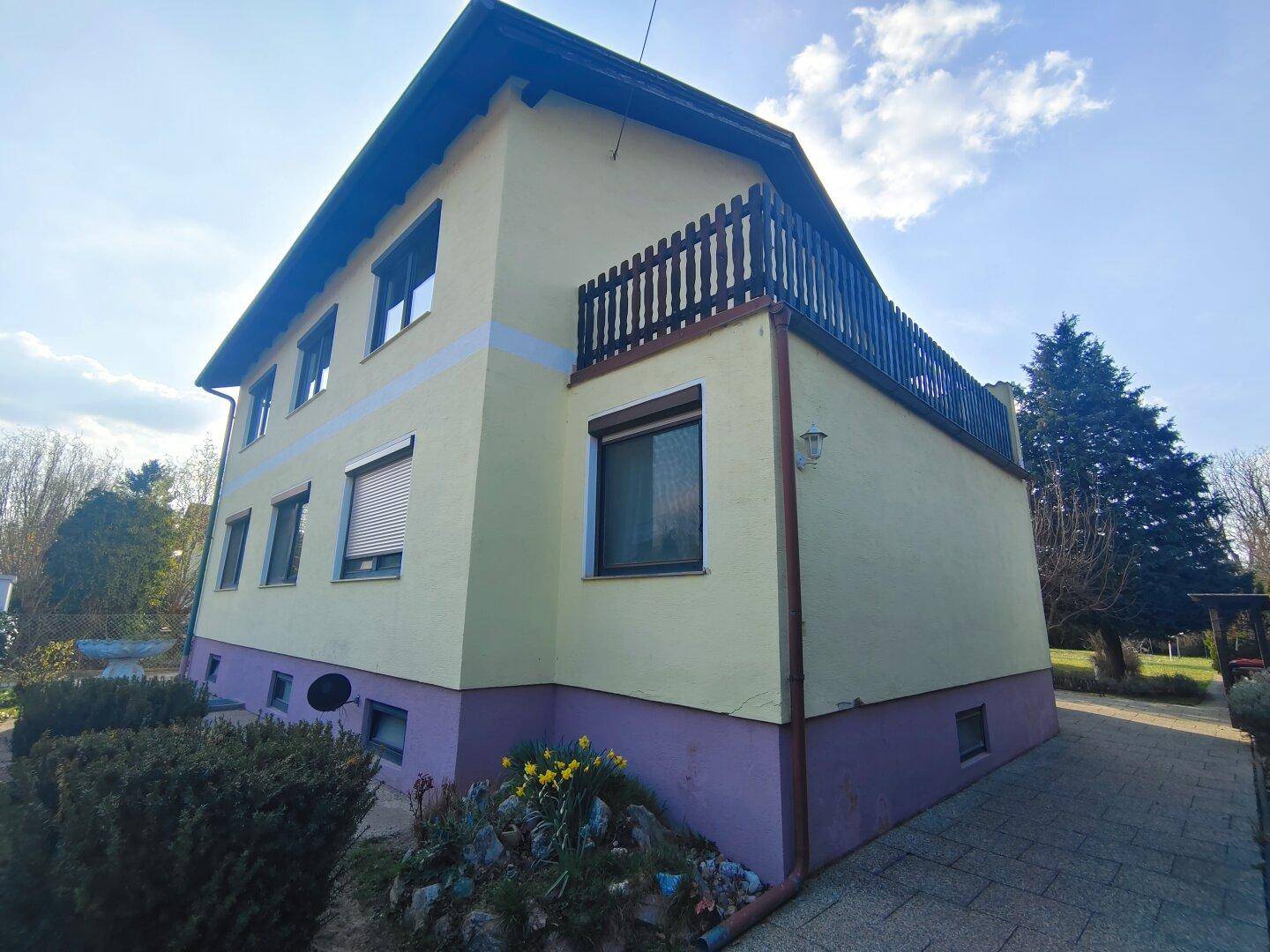 Seitenansicht des Hauses mit einem kleinen Balkon, Blumenbeeten und einer gepflasterten Fläche.