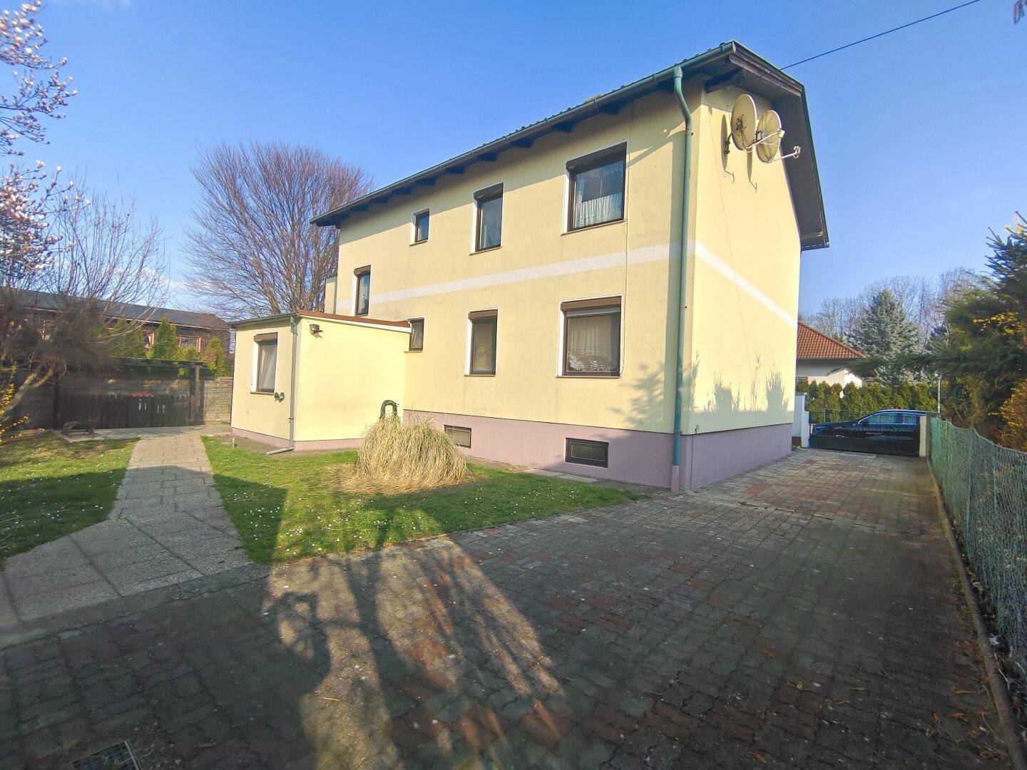 Seitenansicht des Hauses mit gepflasterter Auffahrt, Rasenfläche und einem kleinen Anbau.