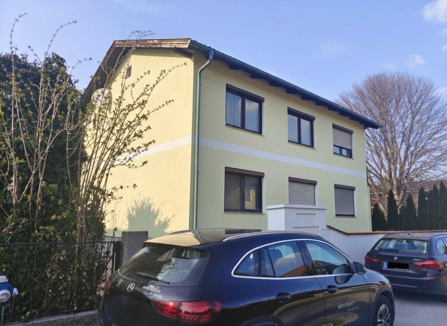 Ansicht des Wohnhauses von der Straße mit gelber Fassade, dunklen Fenstern und geparkten Autos.