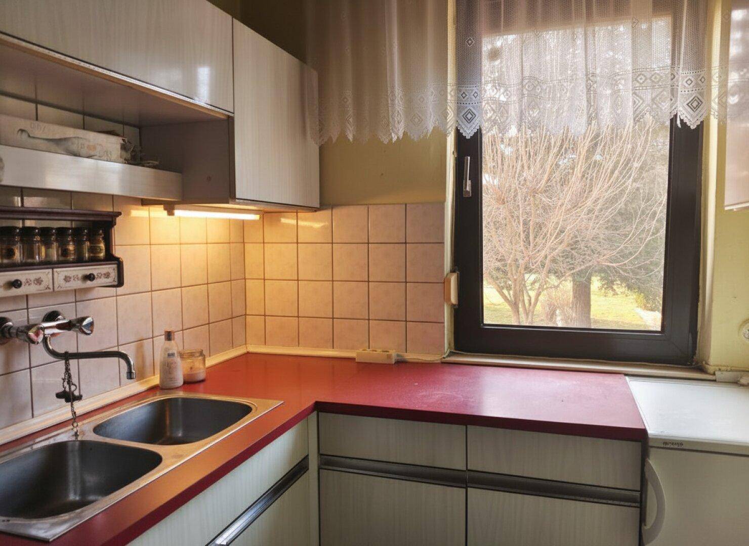 Küche mit weißen Oberschränken, roter Arbeitsplatte und einem Fenster mit Blick ins Grüne.