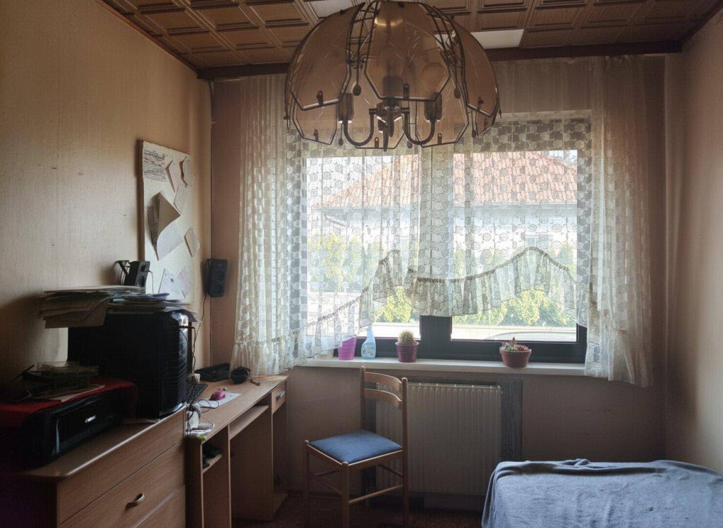 Schlafzimmer oder Arbeitszimmer mit Fenster, Holzdecke und älterer, funktionaler Einrichtung.