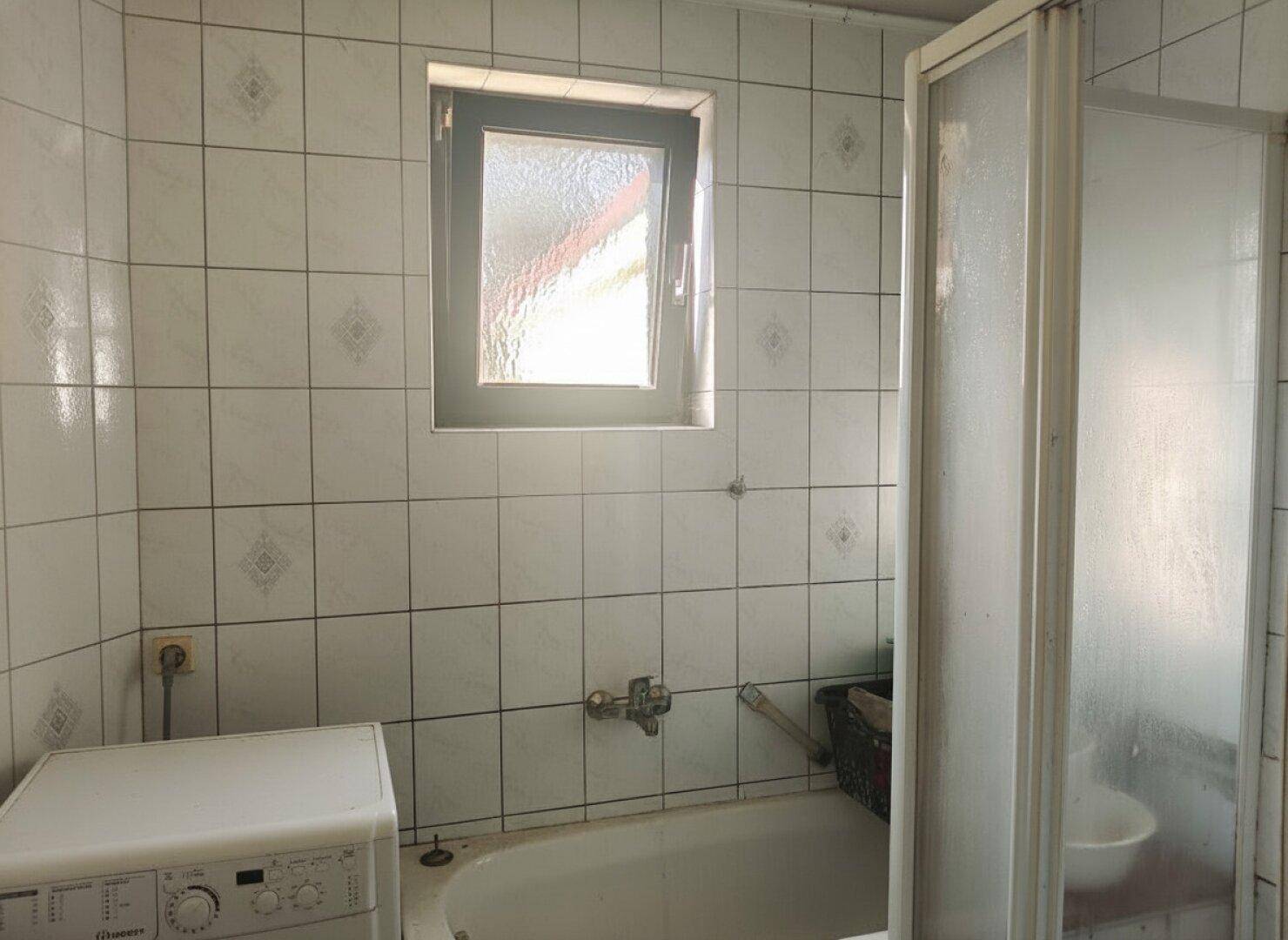 Badezimmer mit weißen Fliesen, Badewanne, Duschkabine und einem Fenster für Tageslicht.