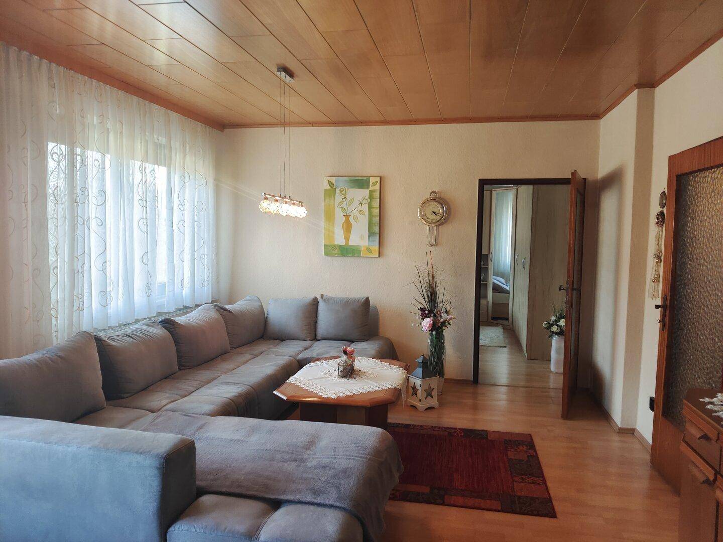 Gemütliches Wohnzimmer mit großem Ecksofa, Holzdecke und hellen Vorhängen am Fenster.