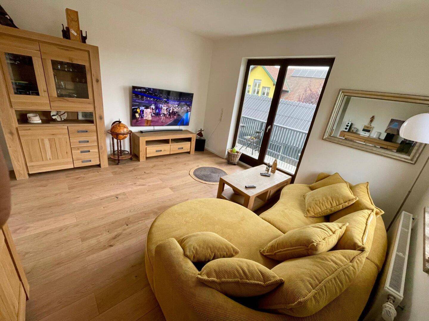 Gemütliches Wohnzimmer mit gelbem Sofa, Holzboden und Blick ins Grüne durch große Fenster.