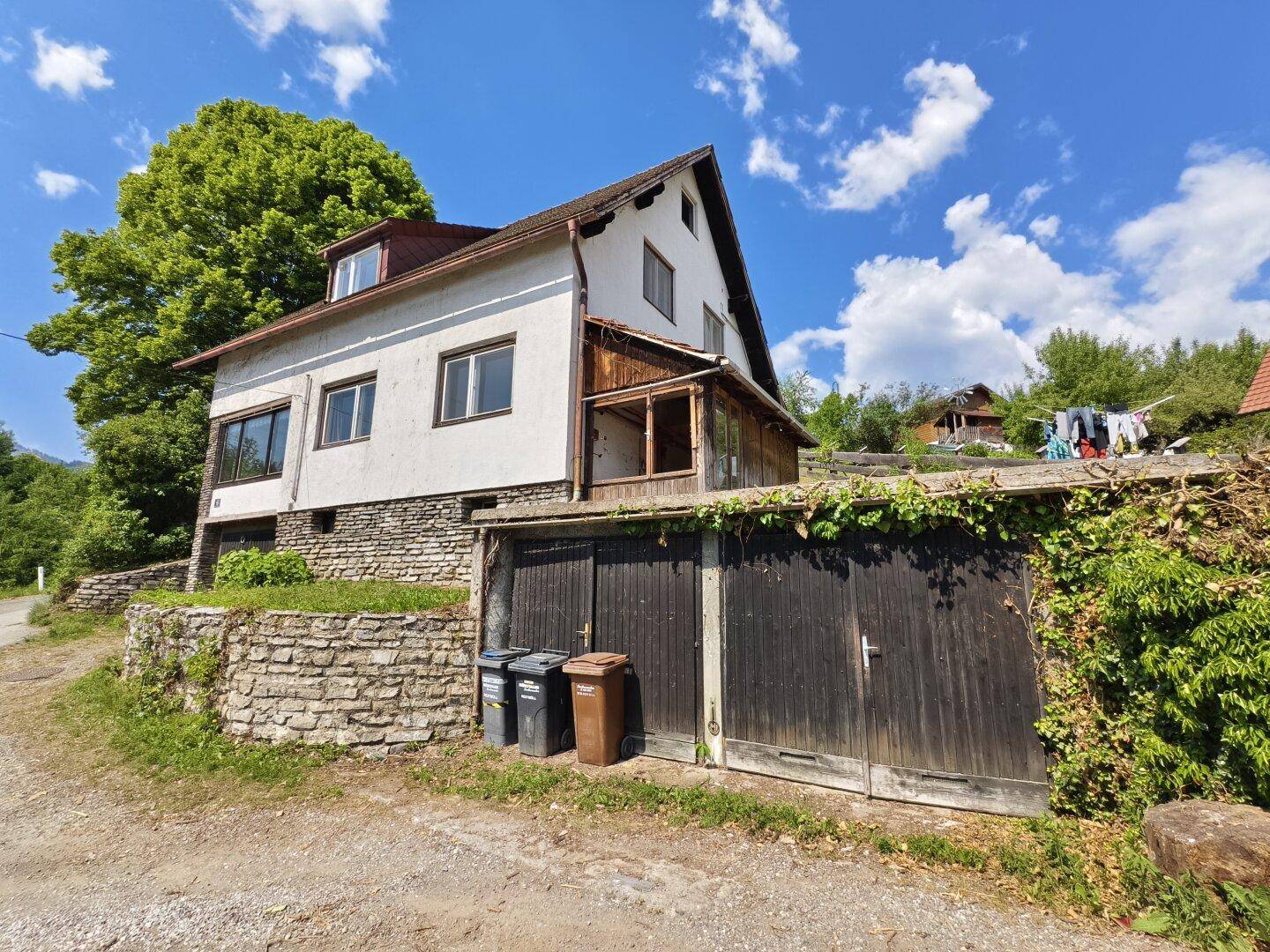 Haus mit Steinfassade und Holztoren, umgeben von üppigem Grün und Bäumen.
