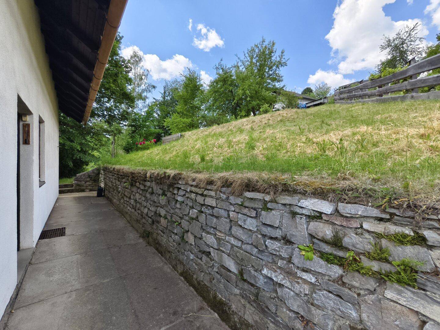 Steingemauerte Wand entlang eines Weges mit Gras und Bäumen im Hintergrund.