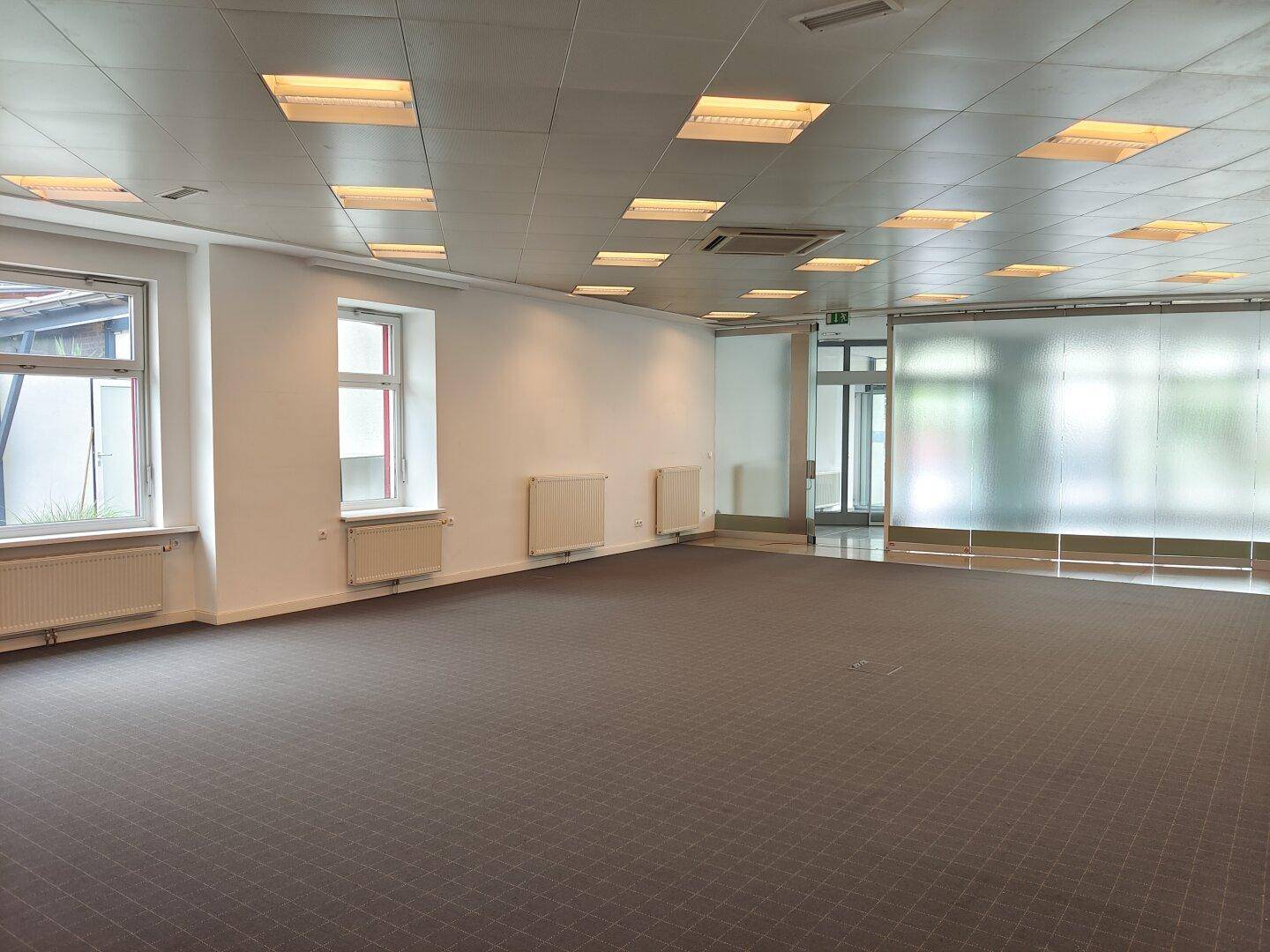 Großer, leerer Raum mit Teppichboden, Rasterdecke und Fenstern für Tageslicht.
