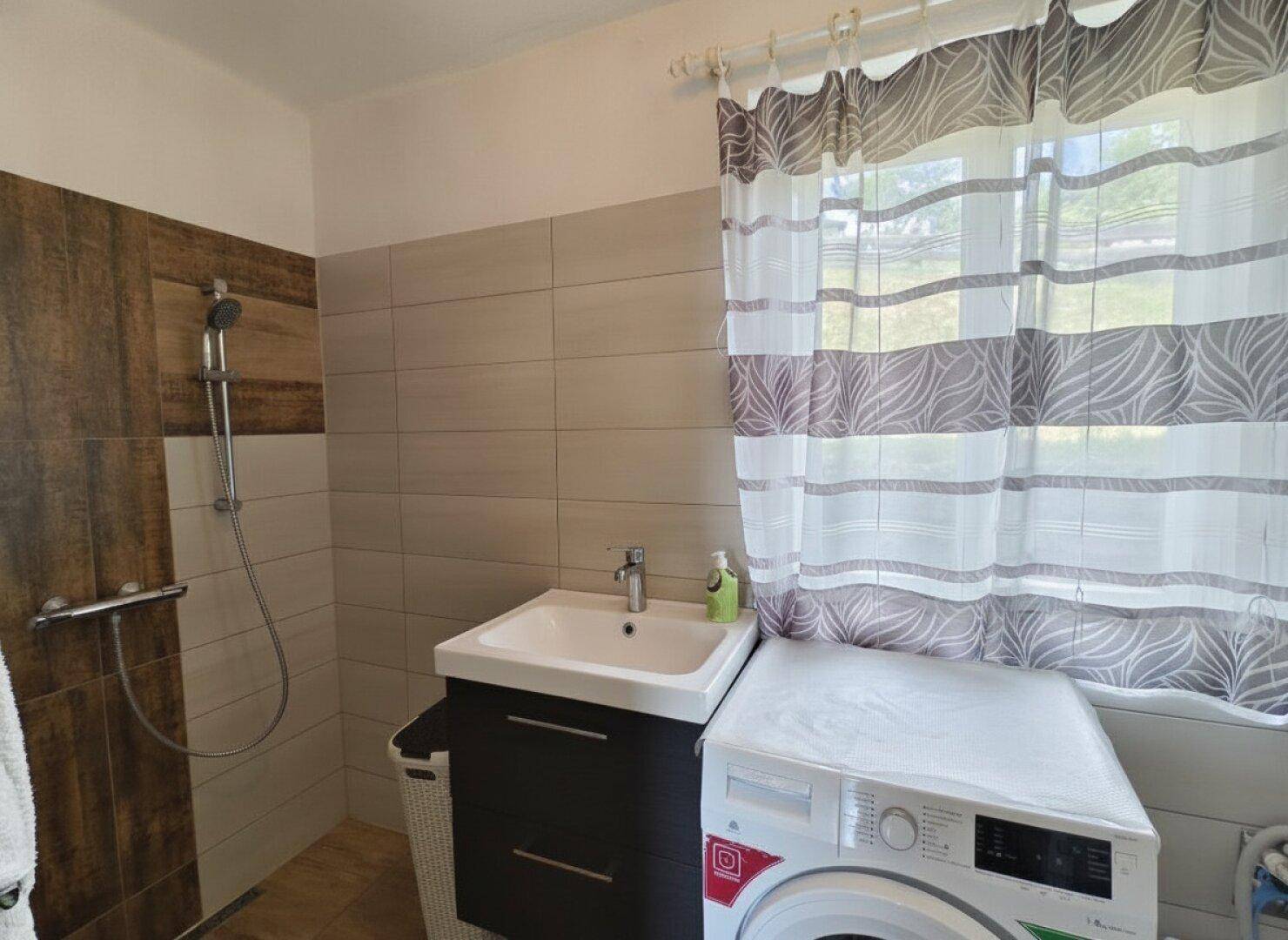 Badezimmer mit Duschbereich, Waschbeckenunterschrank und Waschmaschine am Fenster.
