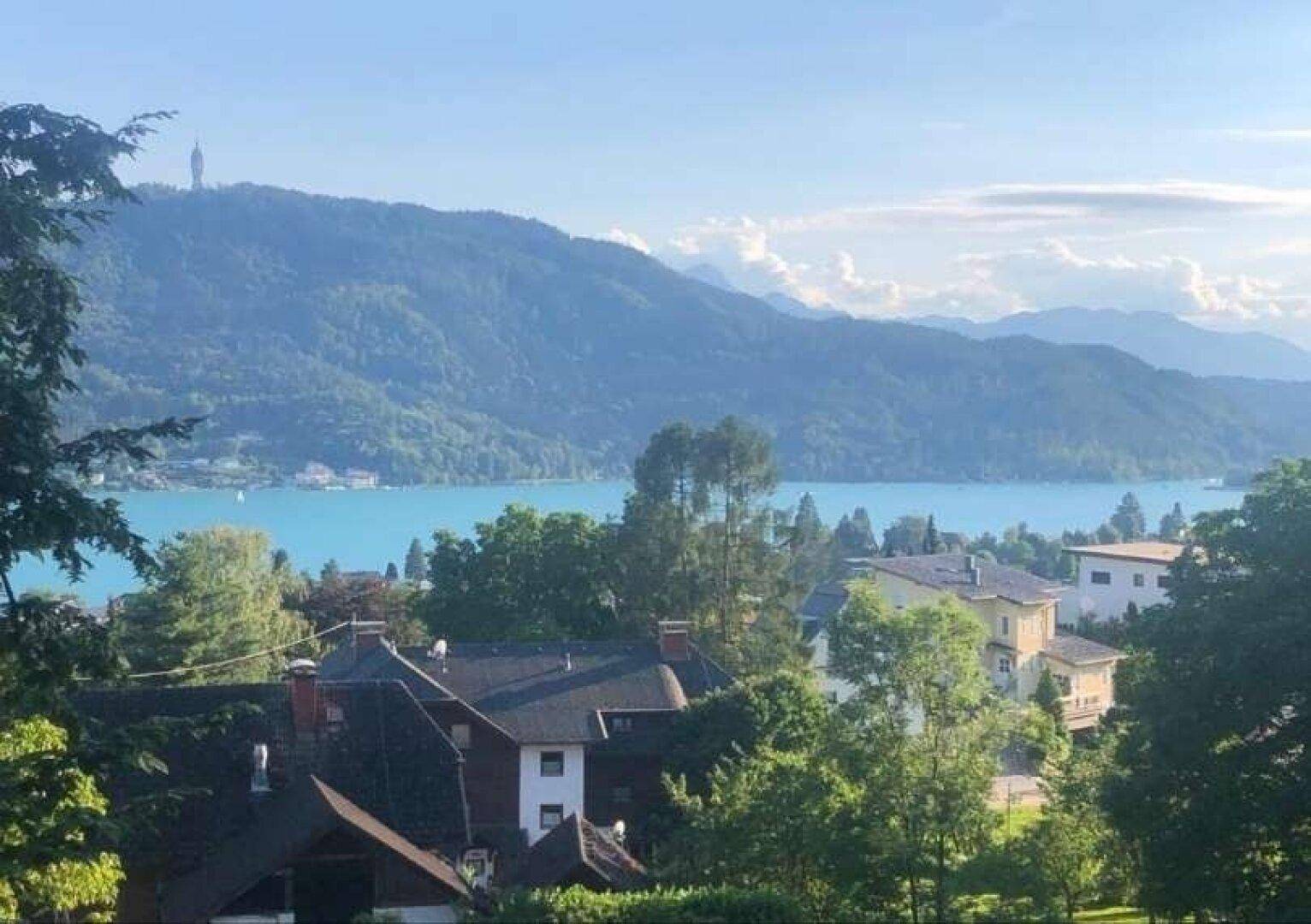 Weitläufiger Blick über einen See und bewaldete Hügel mit vereinzelten Häusern im Vordergrund.
