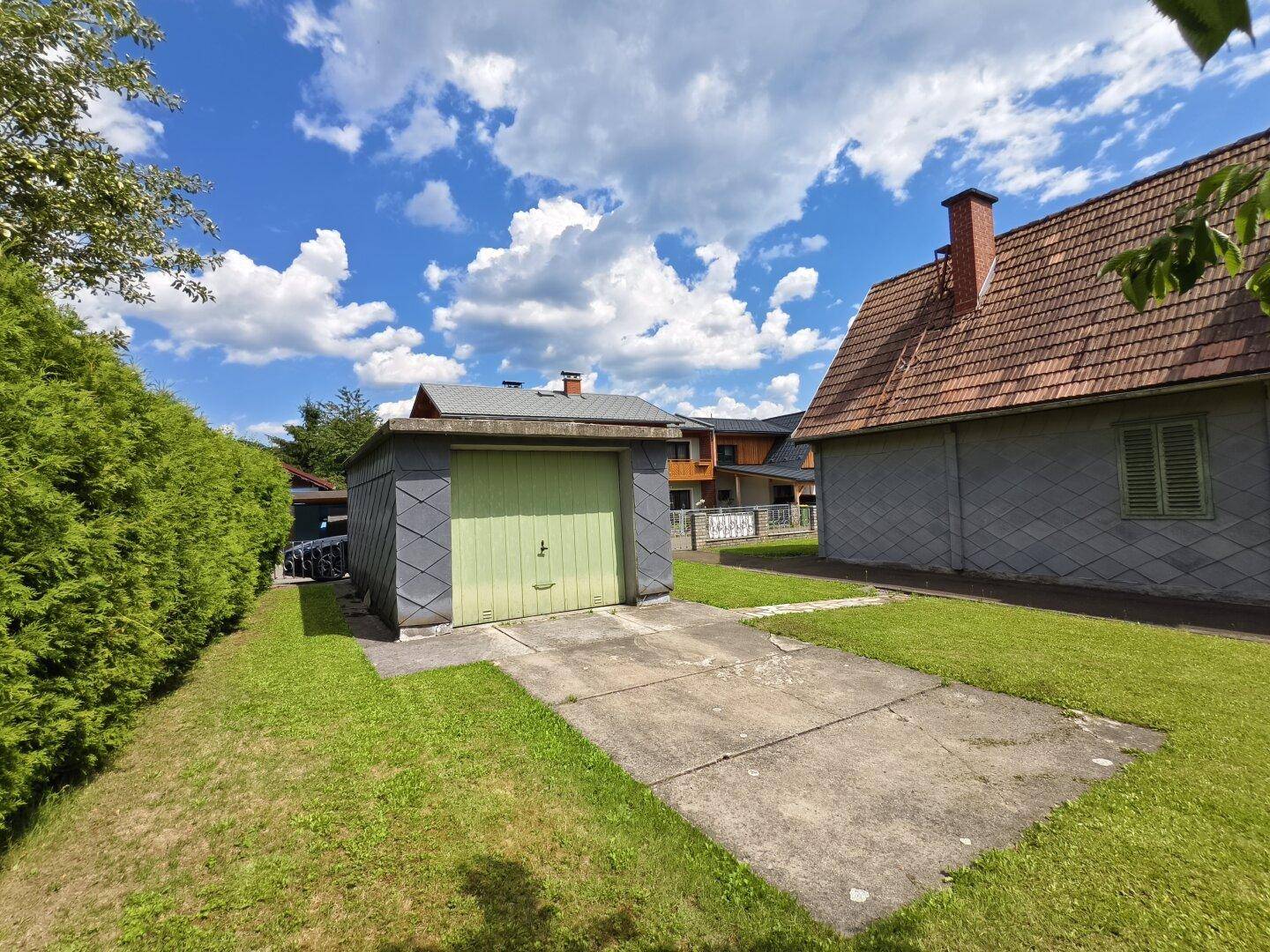 Gartenbereich mit Rasenfläche, Hecke und einer freistehenden Garage unter blauem Himmel.