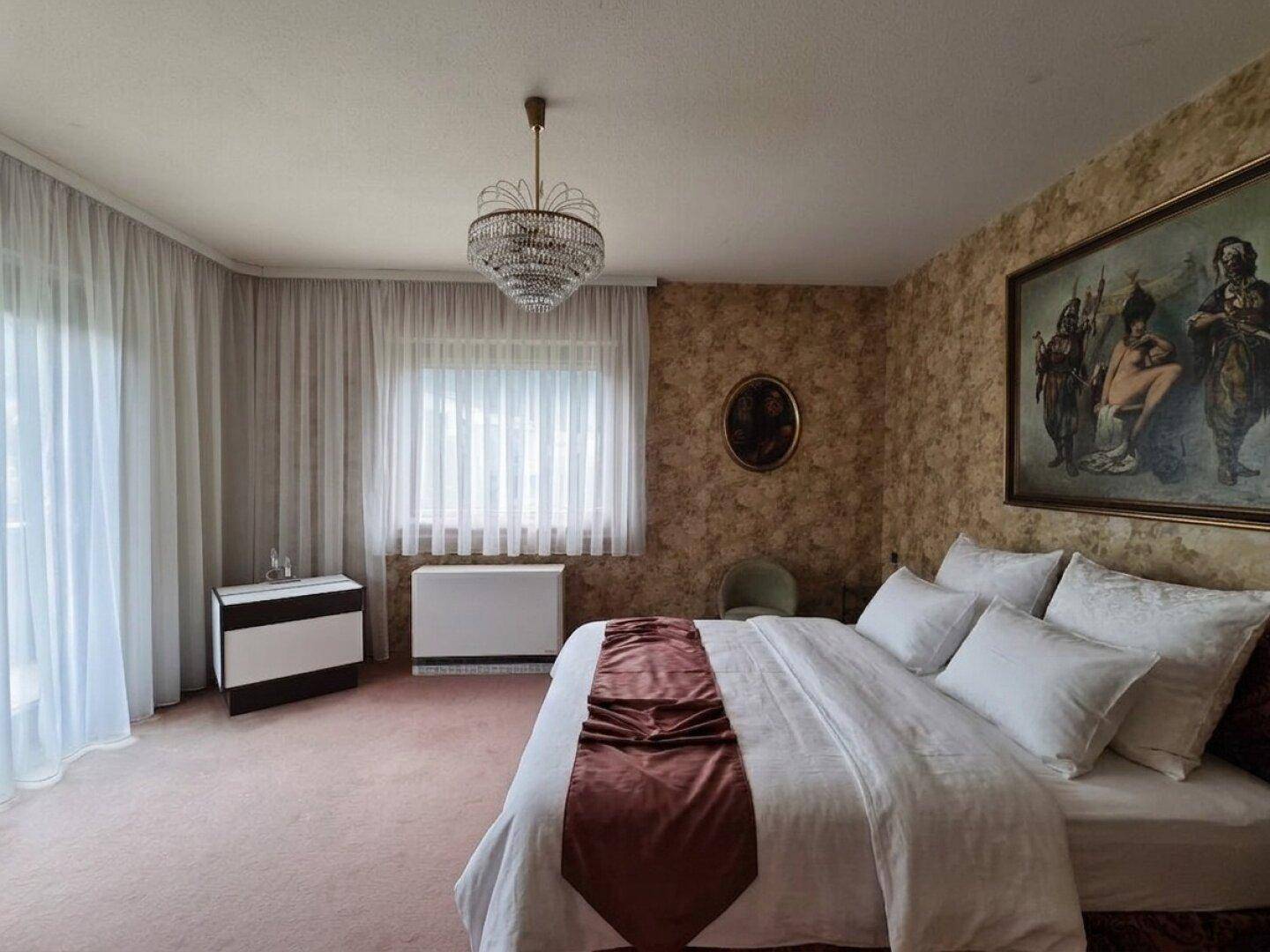 Schlafzimmer mit Doppelbett, Teppichboden, Kronleuchter und Fenstern mit Vorhängen.