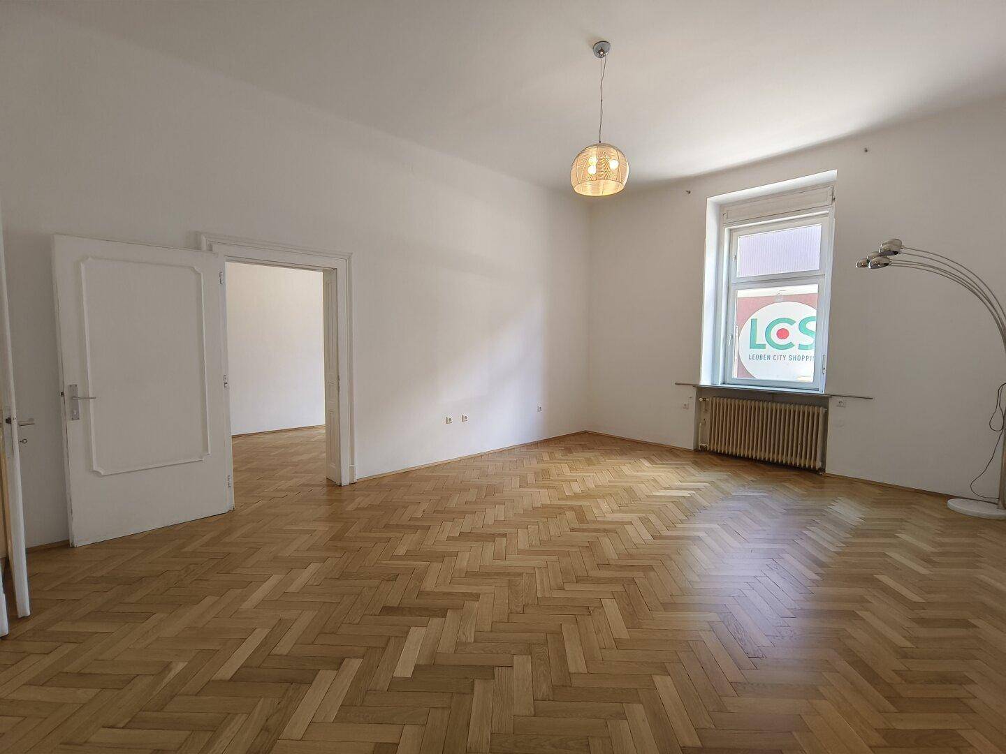 Heller Raum mit Fischgrätparkett, hohen Decken und einem Fenster mit Blick nach draußen.