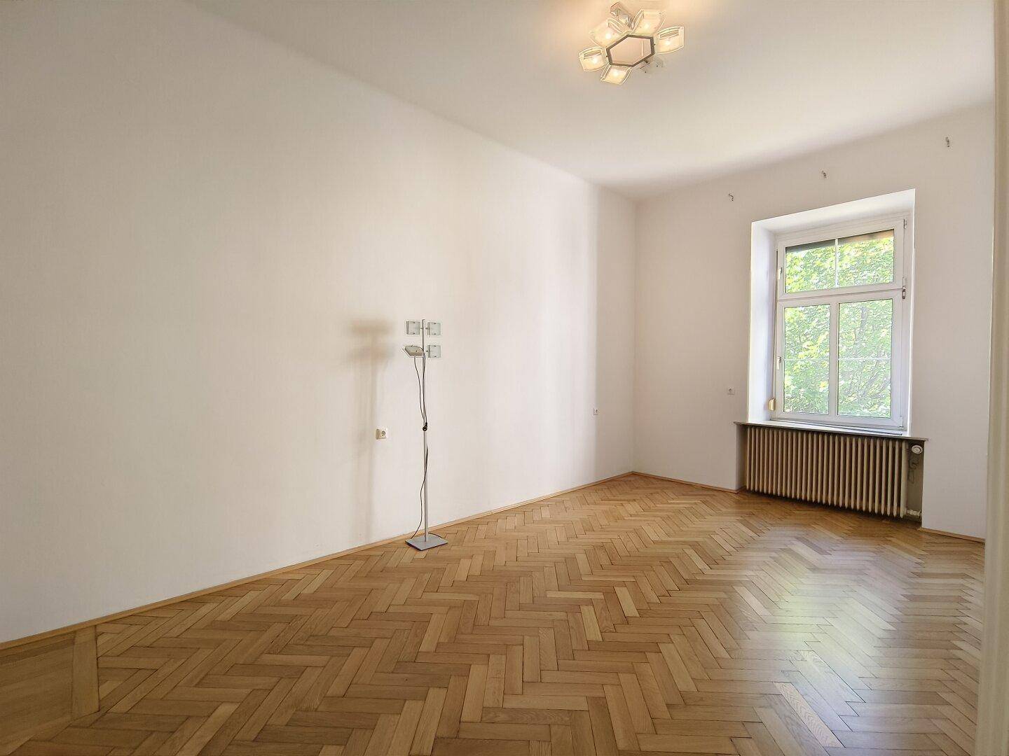 Helles Schlafzimmer mit Fischgrätparkett, großem Fenster und einem Heizkörper unter dem Fenster.