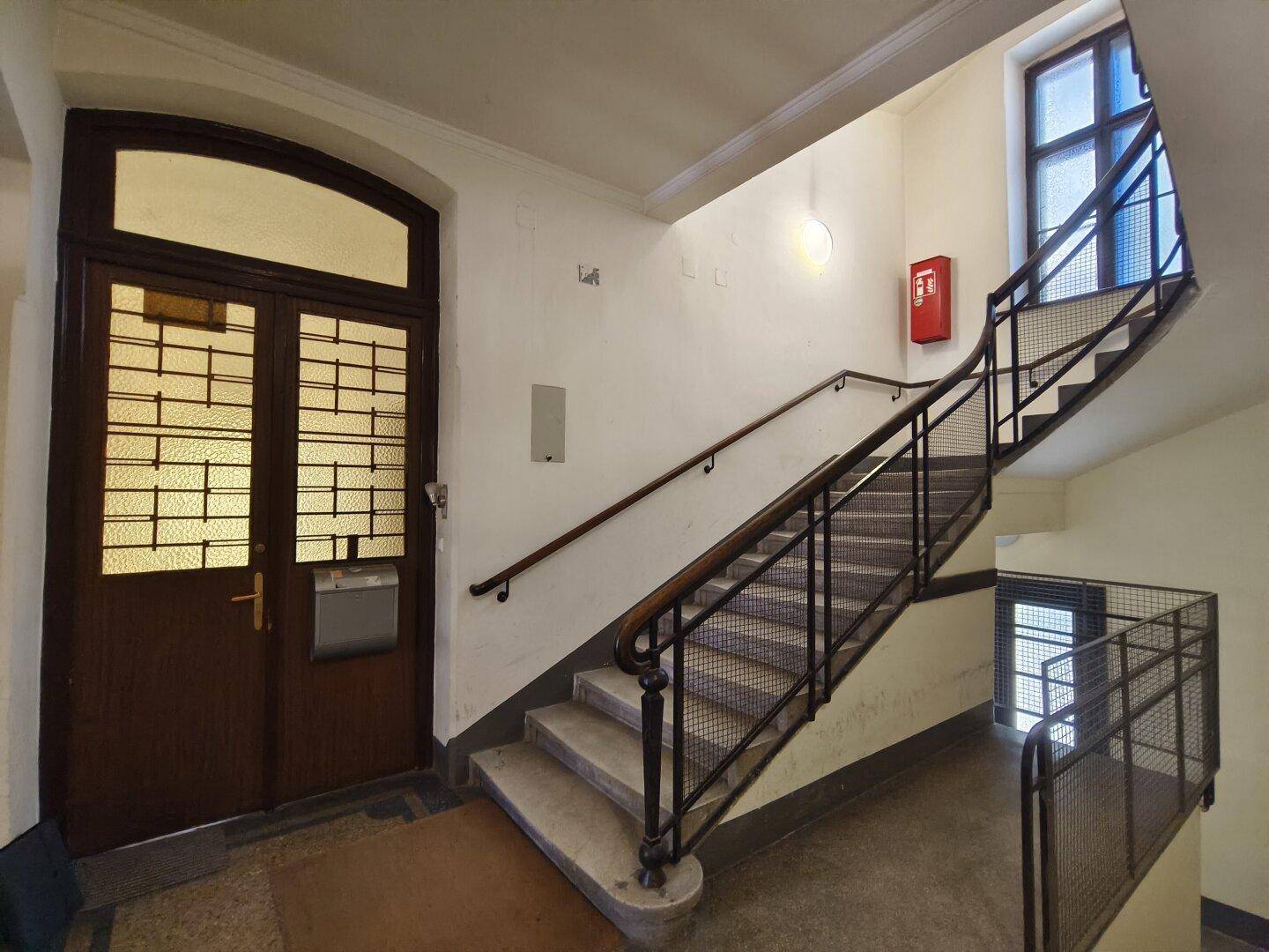 Geräumiges Treppenhaus mit Holzgeländer, Metallstreben und einer großen Eingangstür.