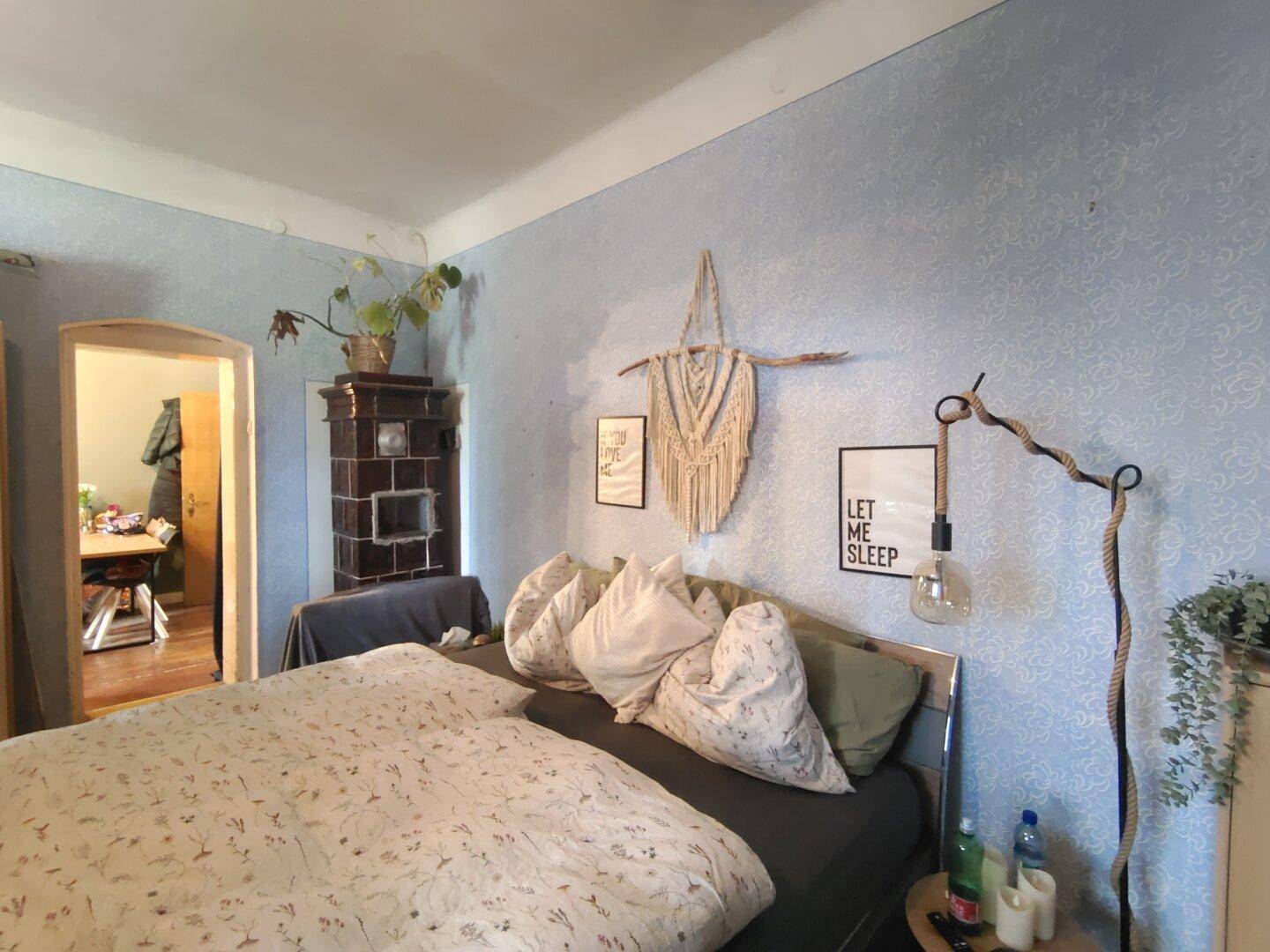 Schlafzimmer mit Bett, einem traditionellen Kachelofen und hellblauer gemusterter Tapete.