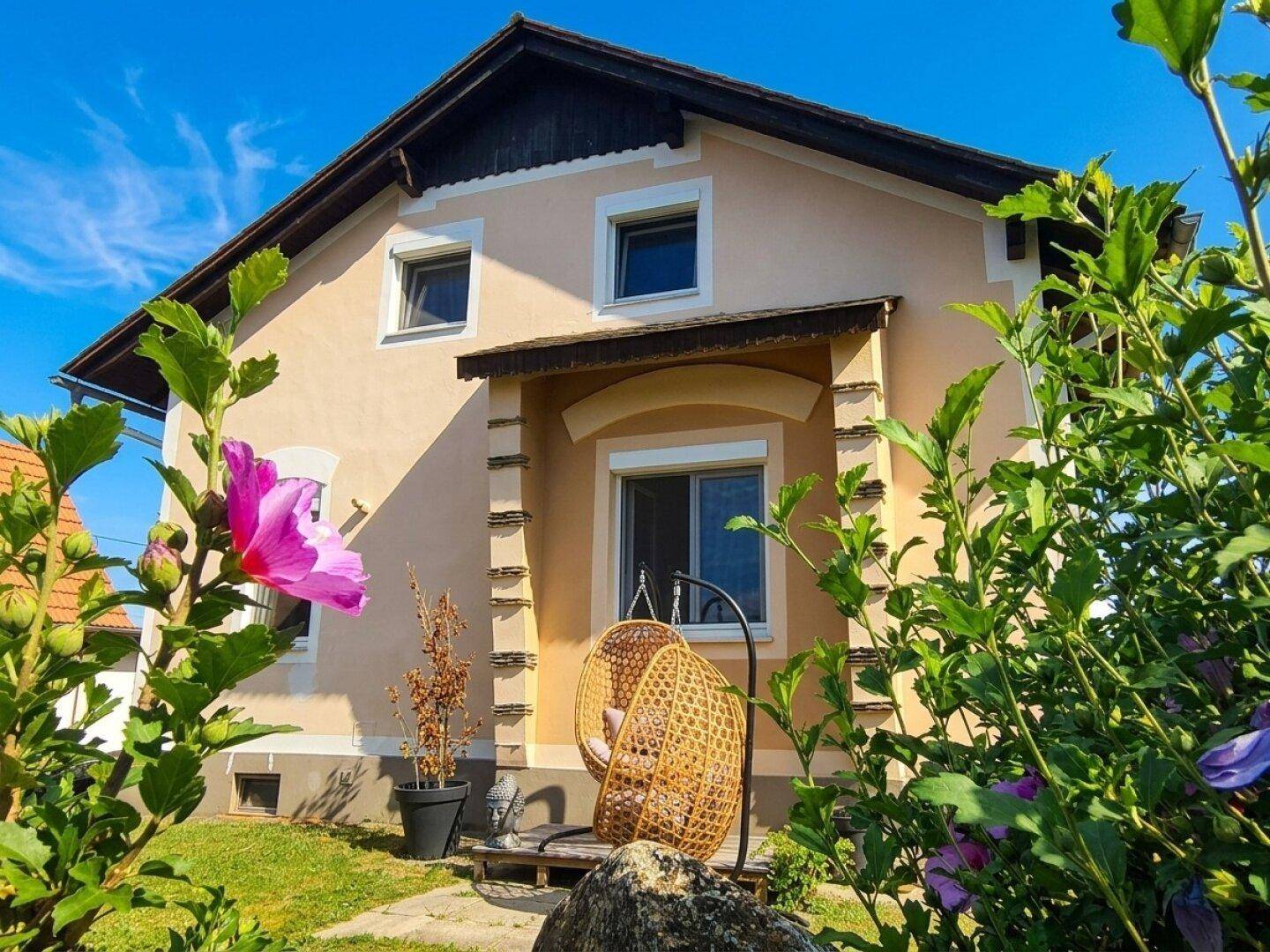 Vorderansicht des Hauses mit kleiner Veranda und einem hängenden Korbsessel im Vorgarten mit blühenden Pflanzen.