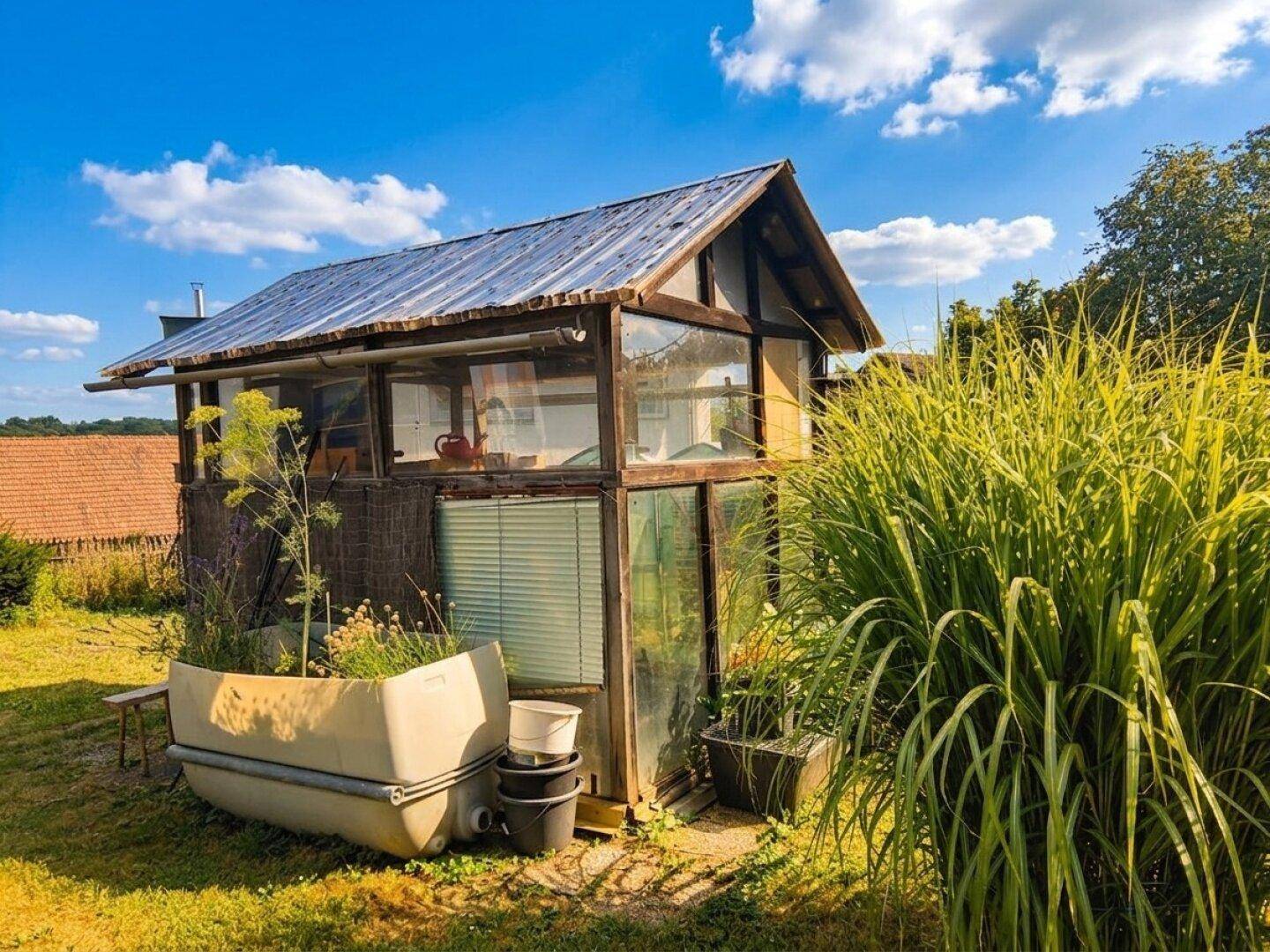 Rustikales Holzgebäude mit Glaswänden, umgeben von üppigem Grün im Gartenbereich unter blauem Himmel.
