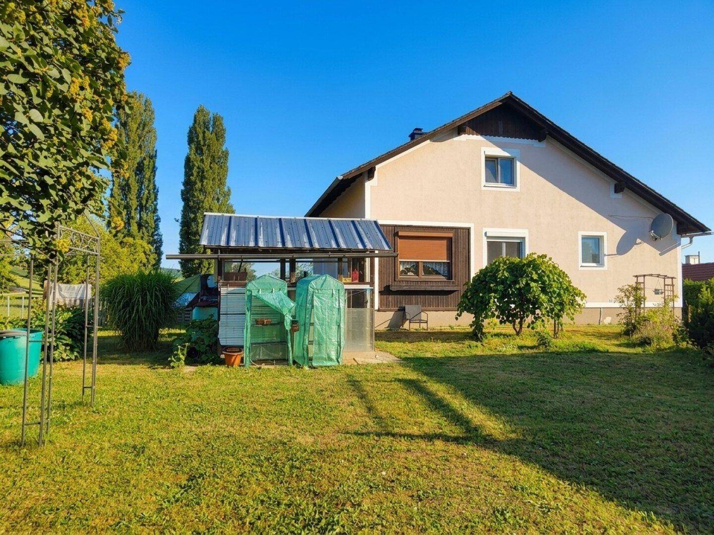 Rückseite des Hauses mit großem Garten und einem kleinen Gewächshaus oder überdachtem Bereich im Sonnenlicht.