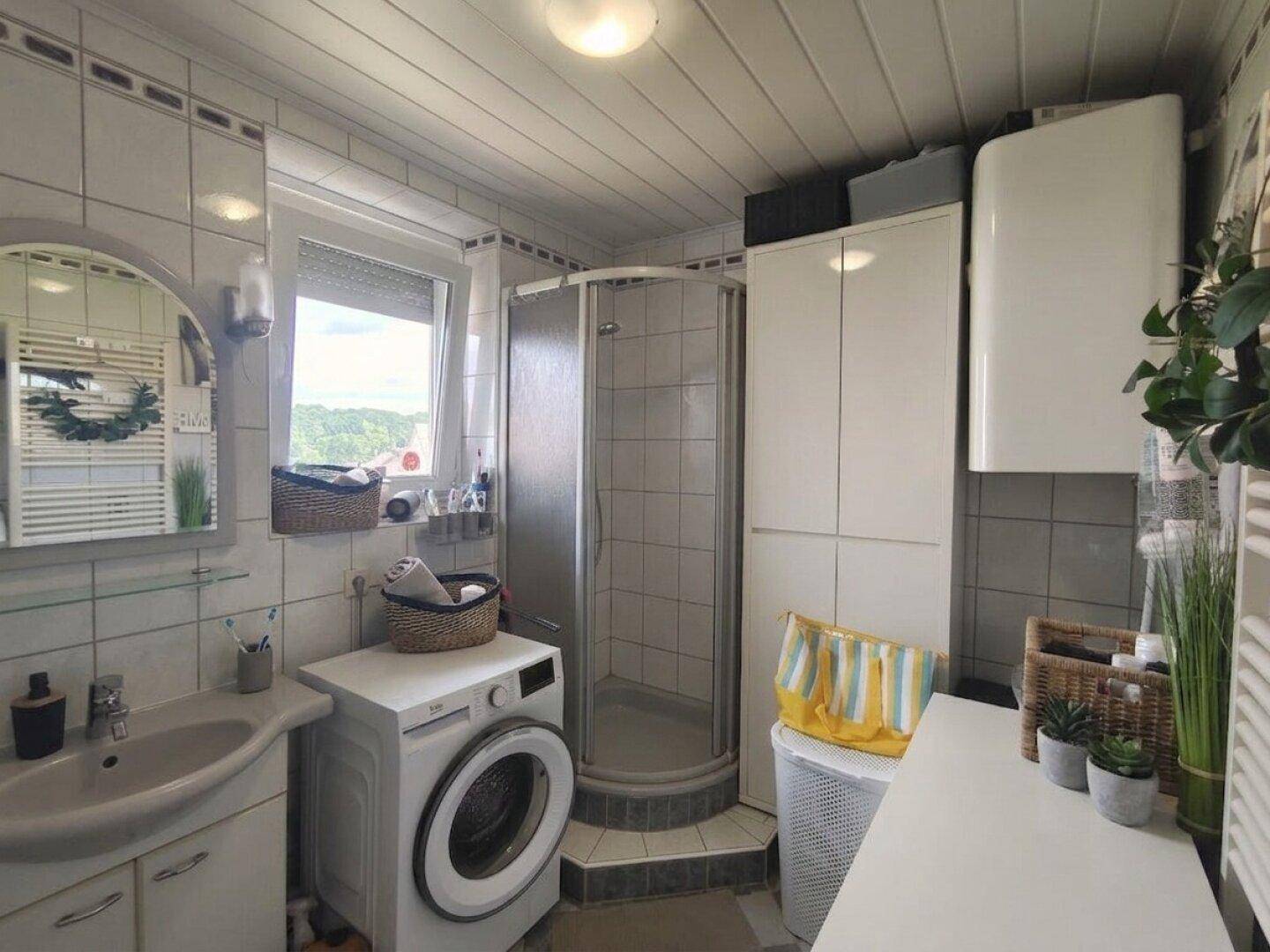 Helles Badezimmer mit Duschkabine, Waschbecken, Waschmaschine und einem Fenster mit Ausblick.