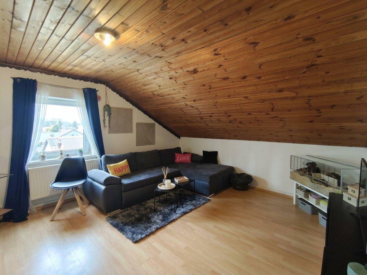 Dachwohnzimmer mit schräger Holzdecke, gemütlichem Sofa und einem Fenster mit Ausblick.
