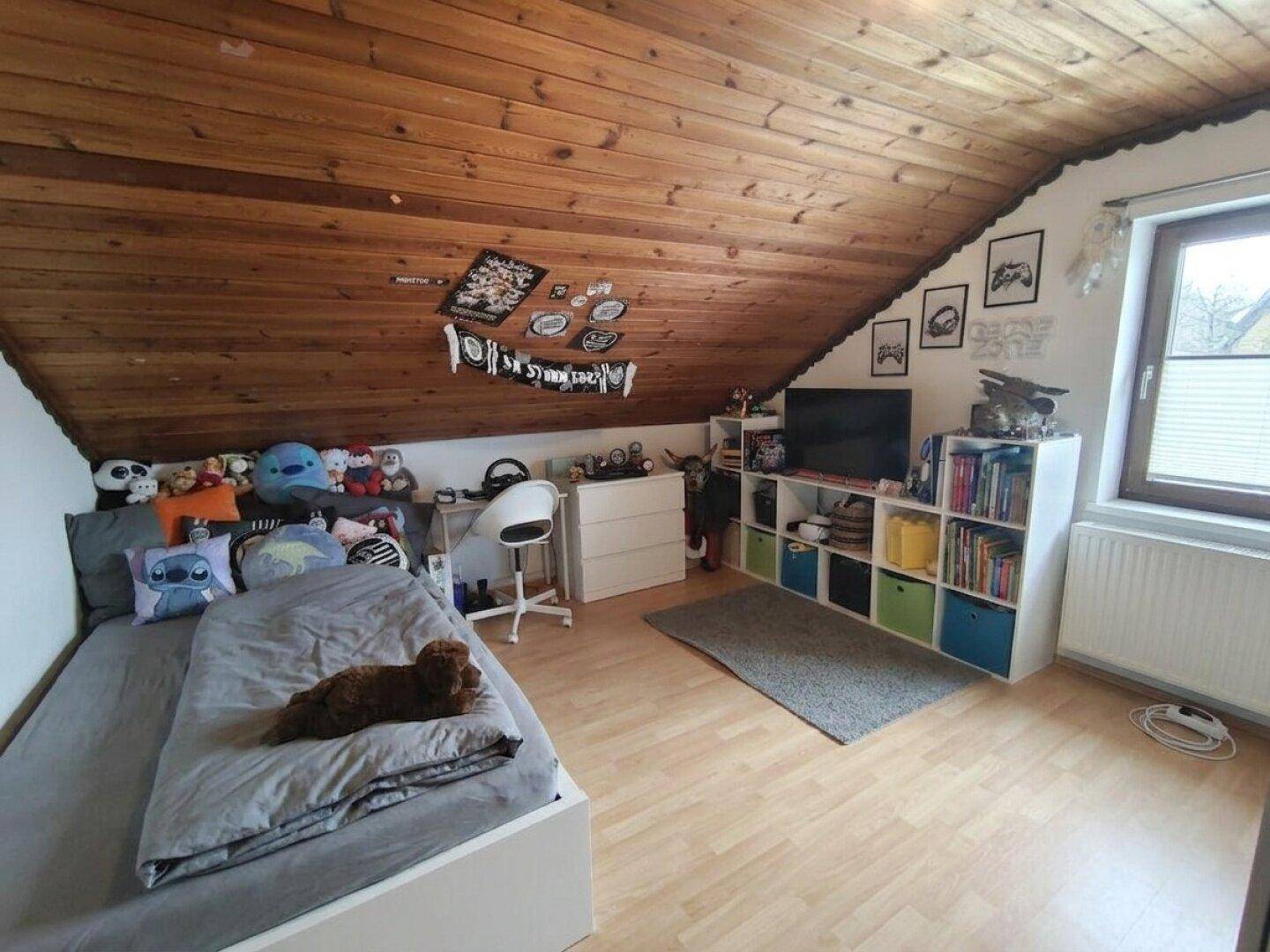 Dachschlafzimmer oder Kinderzimmer mit schräger Holzdecke, Bett und einem Fenster.
