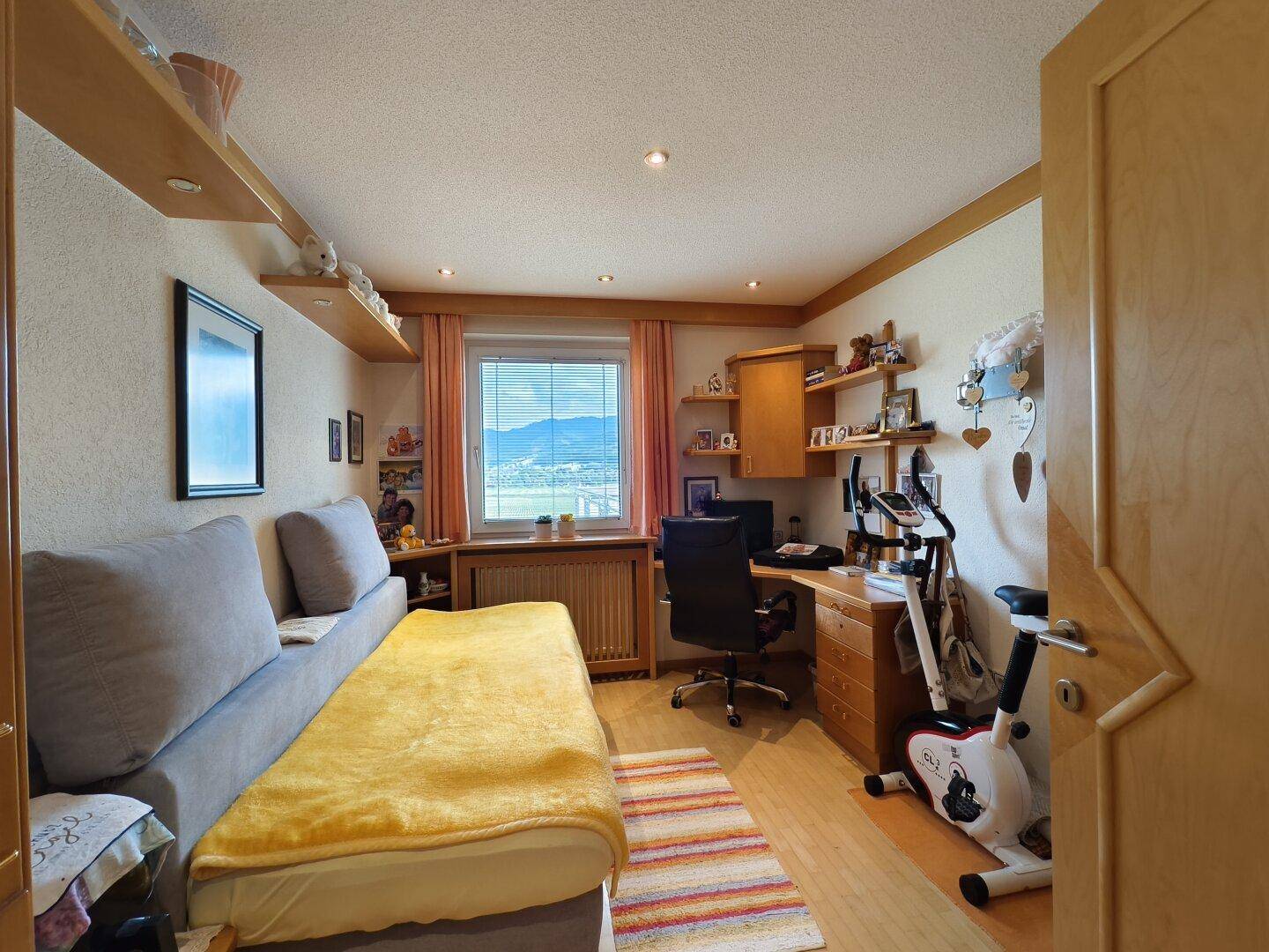 Helles Arbeitszimmer mit Schlafsofa, Schreibtisch und Sportgerät.