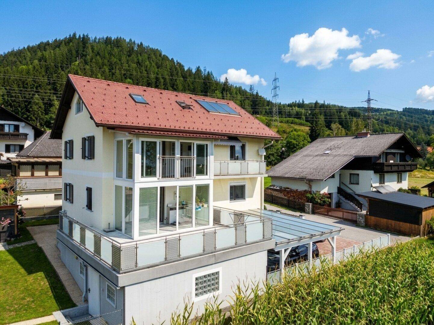Modernes Wohnhaus mit rotem Ziegeldach, großen Glasfronten, Balkonen und Carport in ländlicher Umgebung.
