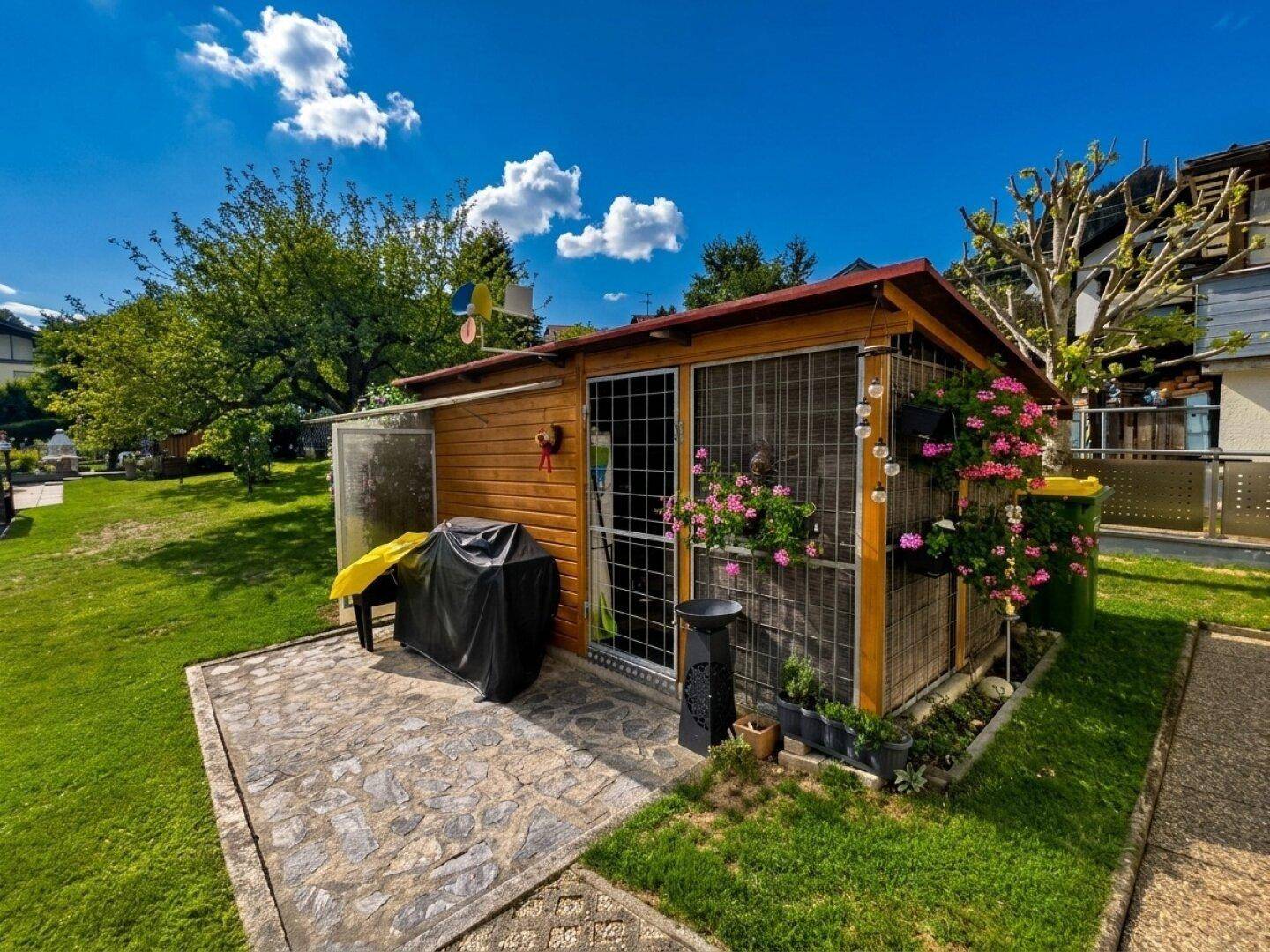 Rustikales Gartenhaus aus Holz mit kleiner Terrasse, Grillbereich und bunten Blumenkästen.
