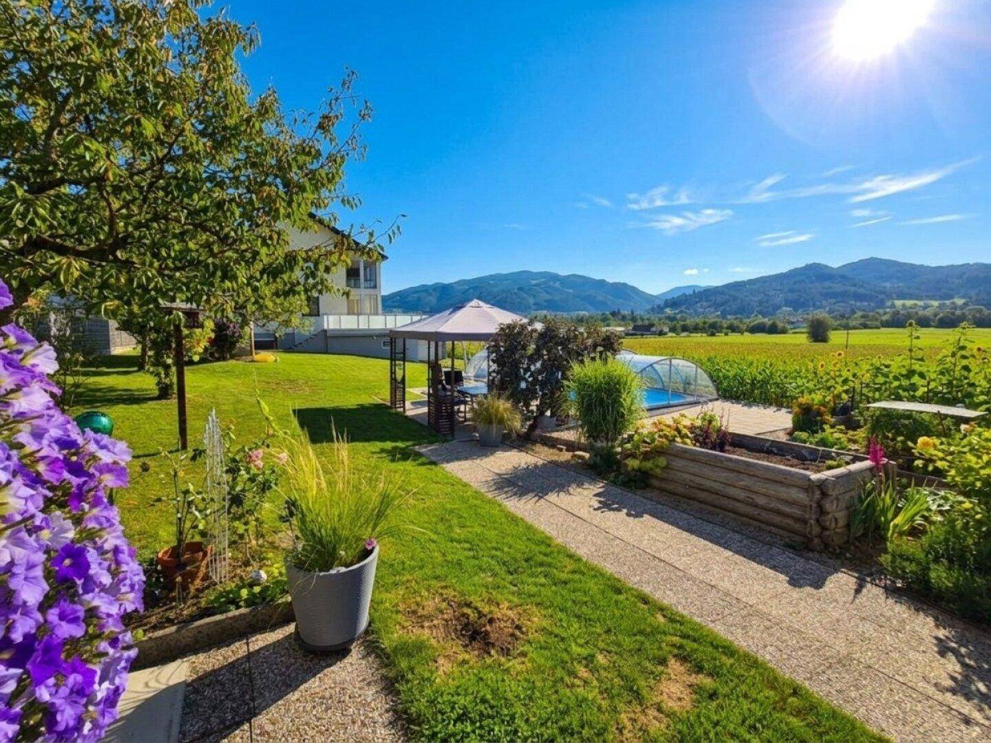 Sonnige Gartenanlage mit Wegen, Hochbeeten und Blick auf das Haus mit Poolbereich.