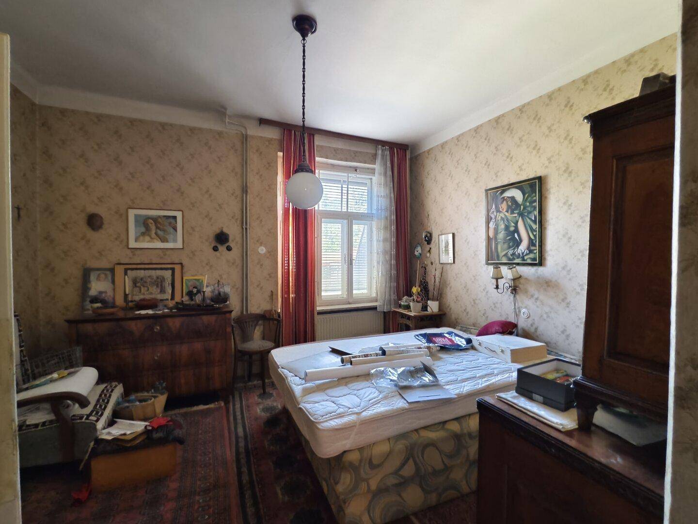 Schlafzimmer mit Bett, Kommode, gemusterter Tapete und Fenster mit Blick nach draußen.