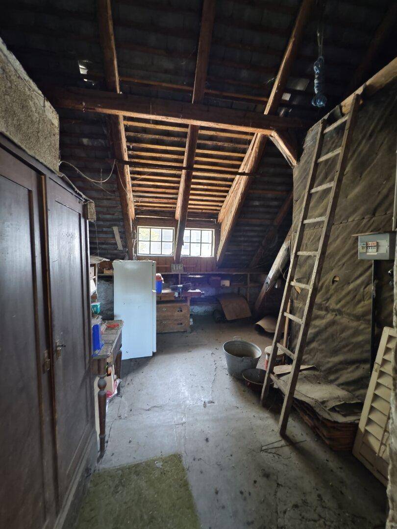 Unausgebauter Dachboden mit sichtbaren Holzbalken, Lagergegenständen und Fenstern.