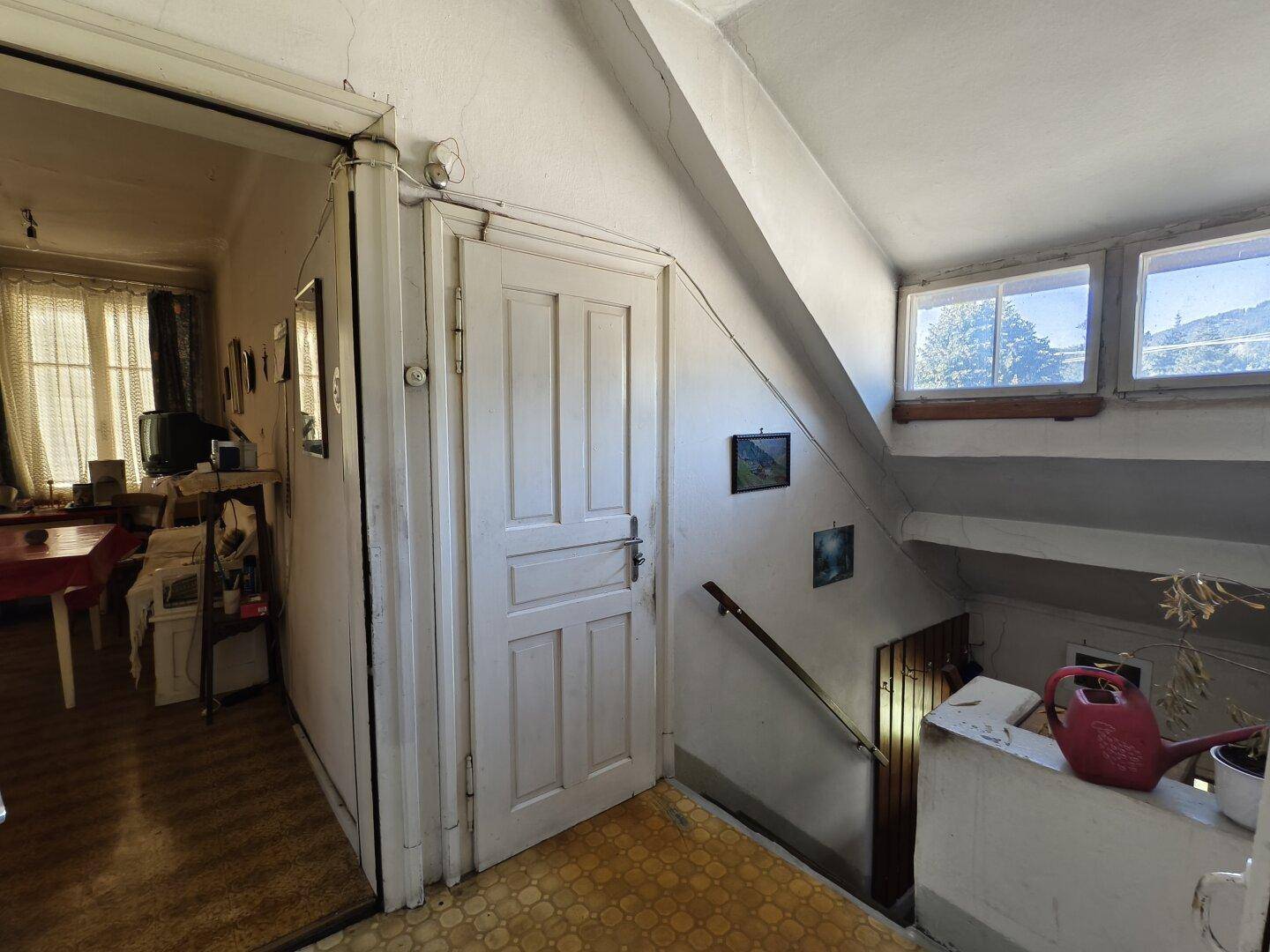 Flur mit Treppenaufgang, Holztür und Fenstern, die Tageslicht hereinlassen.