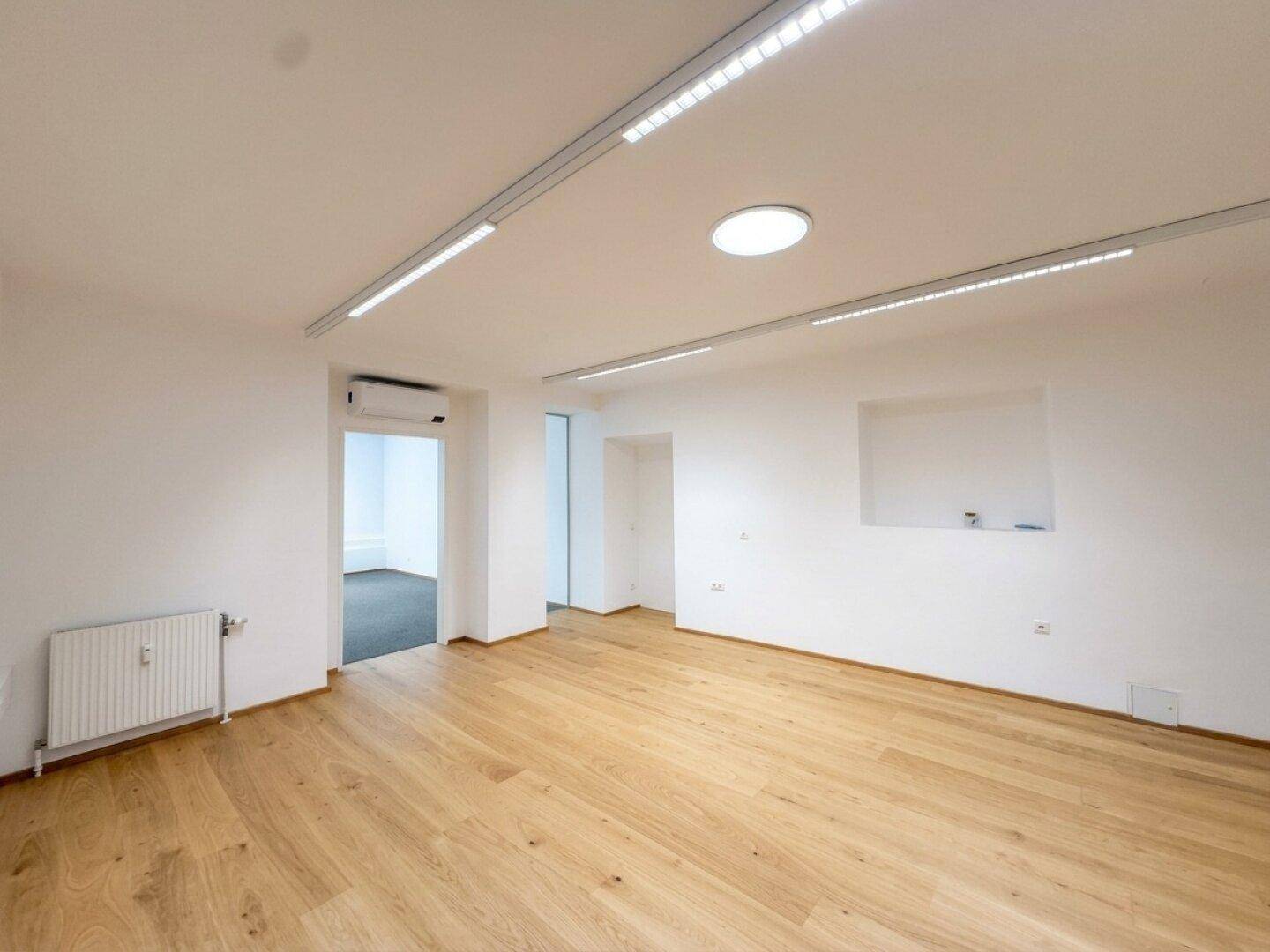 Geräumiger Büroraum mit hellem Holzfußboden, weißen Wänden und moderner Deckenbeleuchtung.