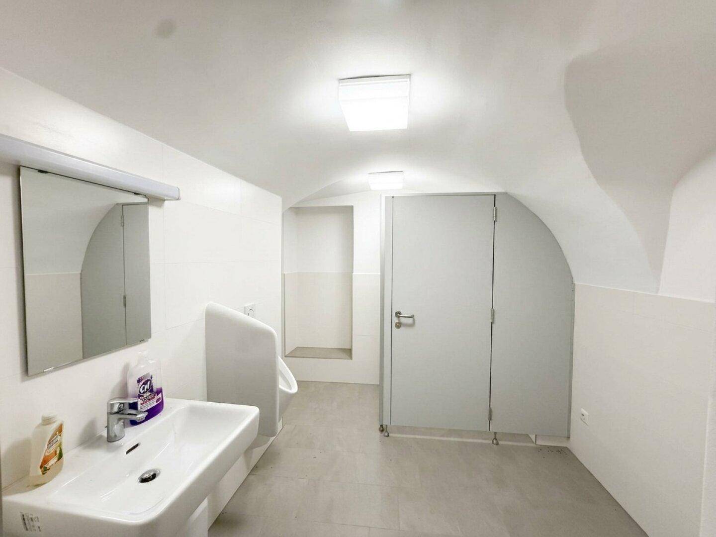 Sauberer öffentlicher Waschraum mit Urinal, Waschbecken und zwei Toilettenkabinen unter einem Gewölbedach.