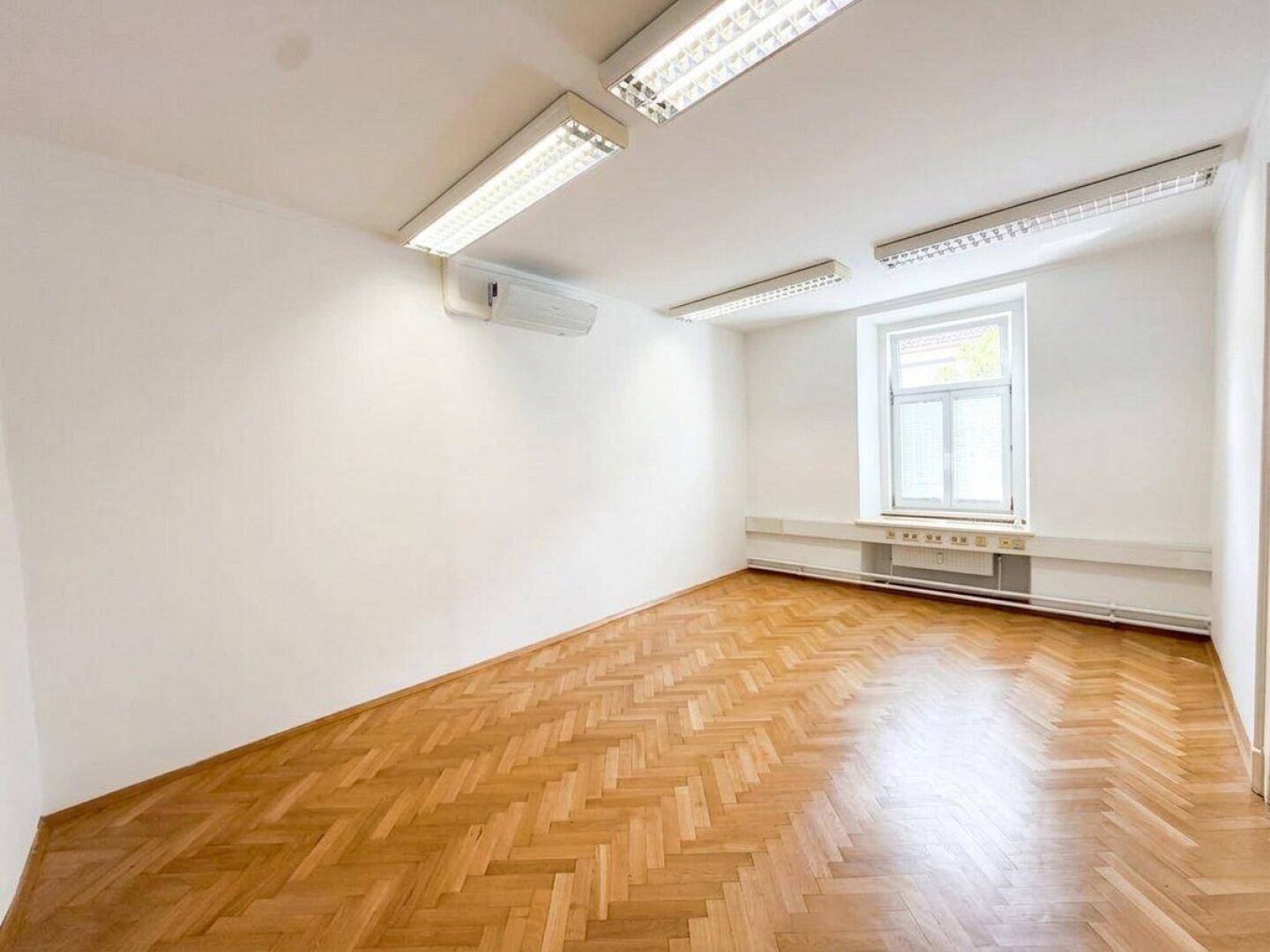 Leerer Raum mit Fischgrätparkettboden, einem Fenster und Klimaanlage an der Wand.