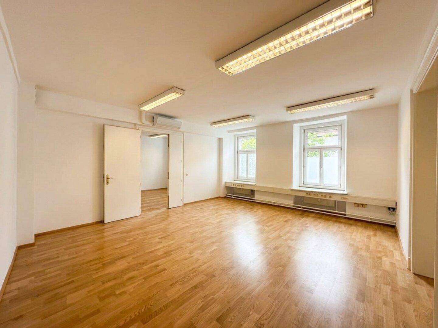 Geräumiger, heller Raum mit Holzboden, zwei Fenstern und Klimaanlage an der Decke.