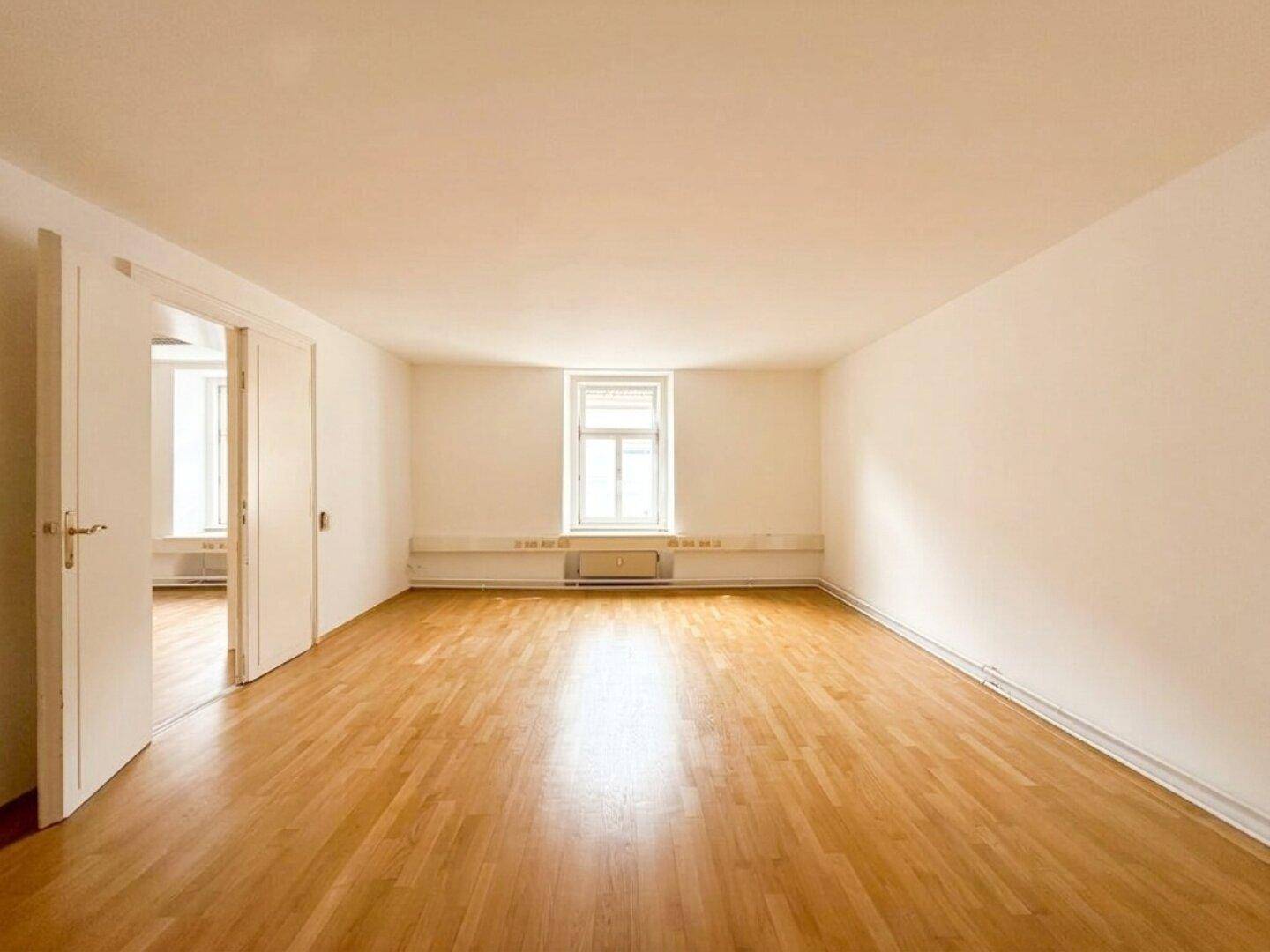 Heller, leerer Raum mit Holzboden und einem großen Fenster, das viel Licht hereinlässt.