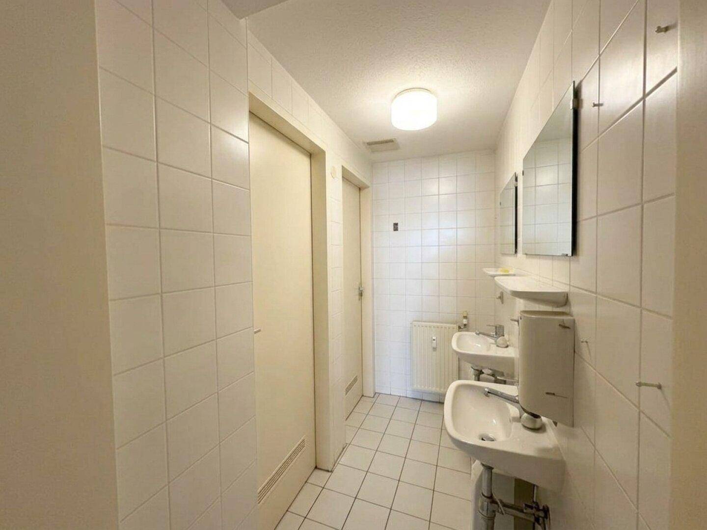 Funktionales Badezimmer mit weißen Fliesen, zwei Waschbecken und einem Heizkörper.